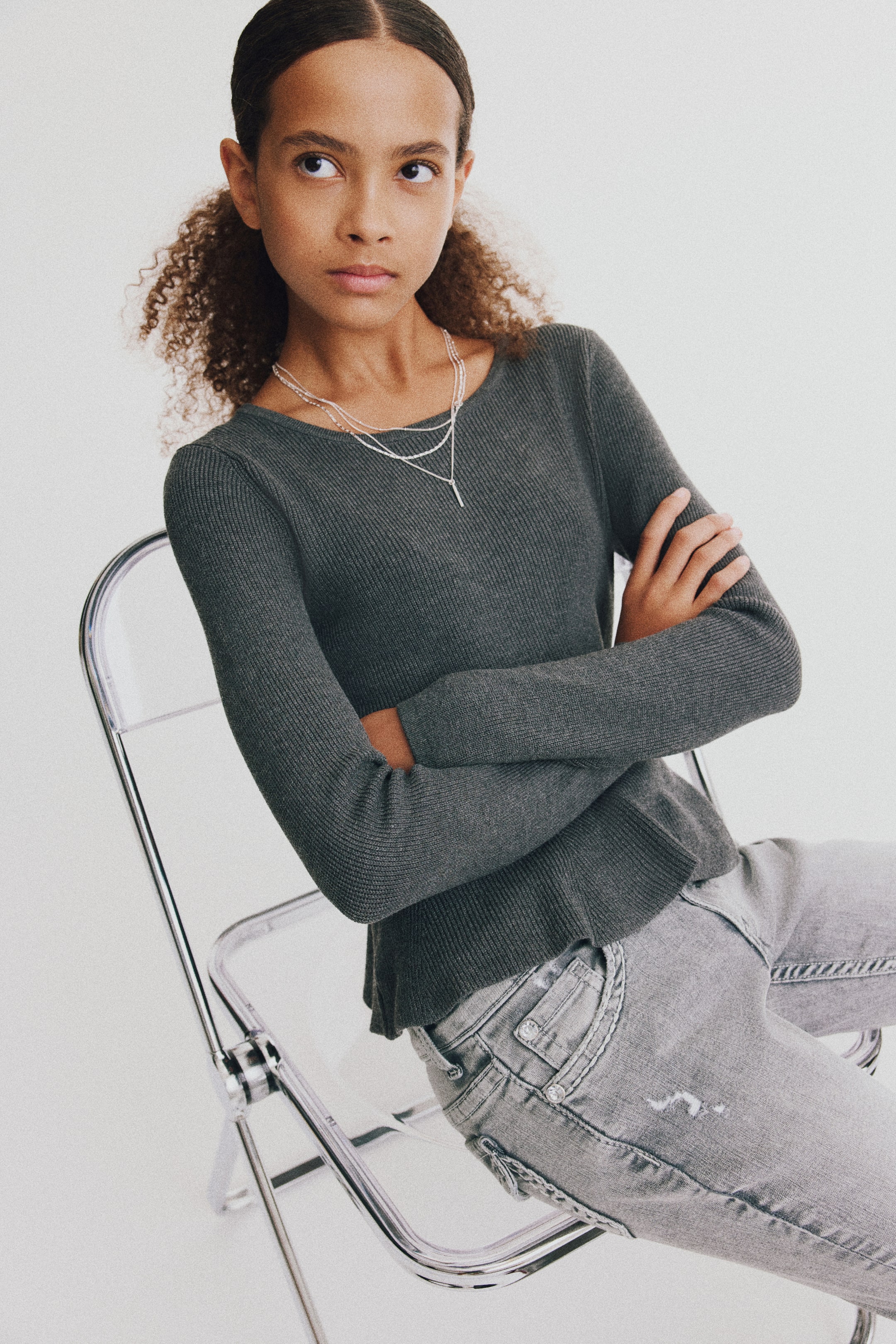 View larger image: Washed gray denim jeans with distressed details are shown from a three-quarter angle on a seated model. The low-waisted jeans feature a front pocket with metallic rivets and a small distressed tear on the thigh.