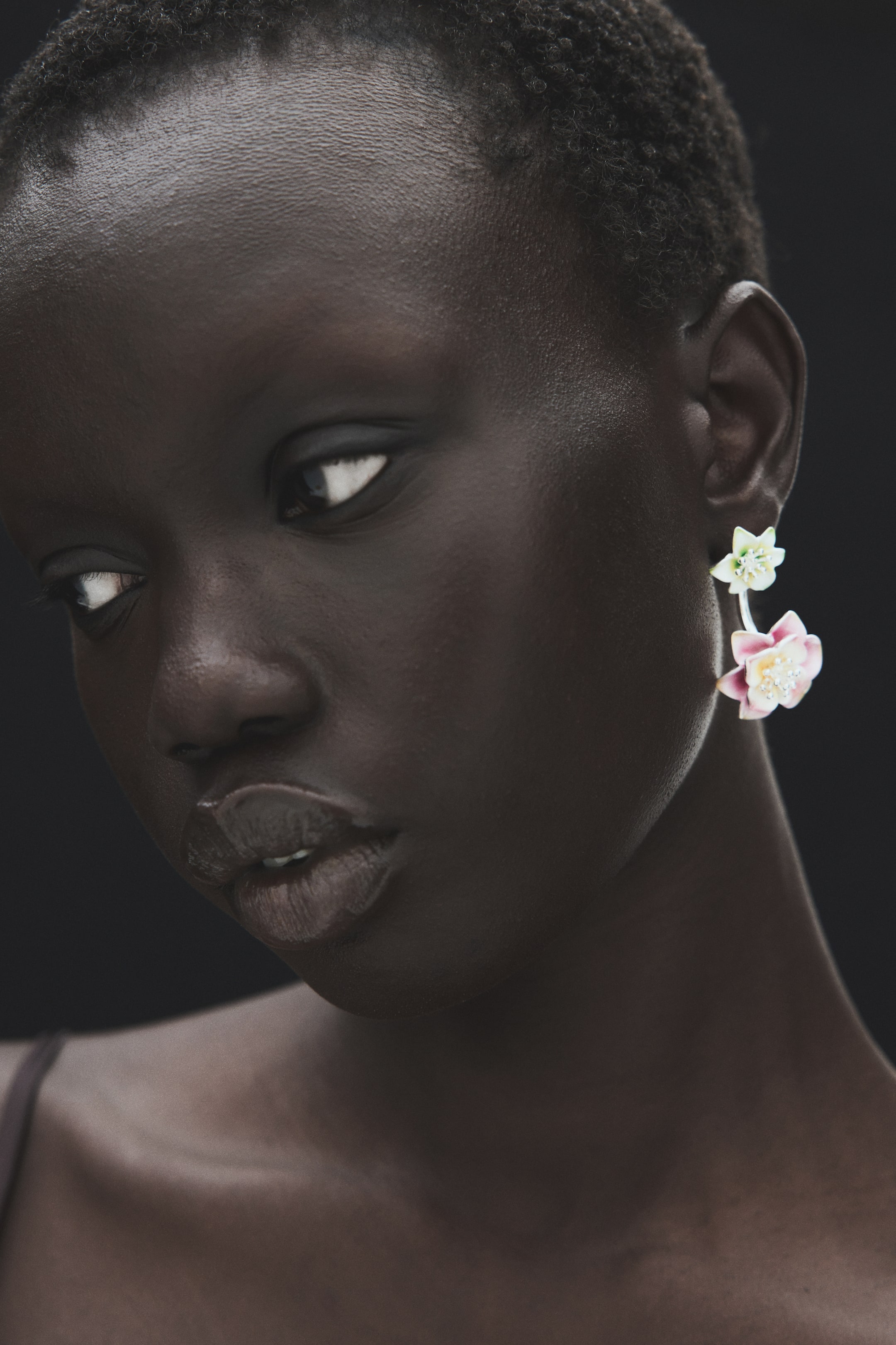 Ampliar la imagen: La modelo lleva aretes metálicos delanteros y traseros con detalles en forma de flor. Una pequeña flor verde claro con detalles brillantes está colocada en el lóbulo de la oreja, y debajo cuelga un colgante más grande en forma de flor rosa con detalles brillantes.