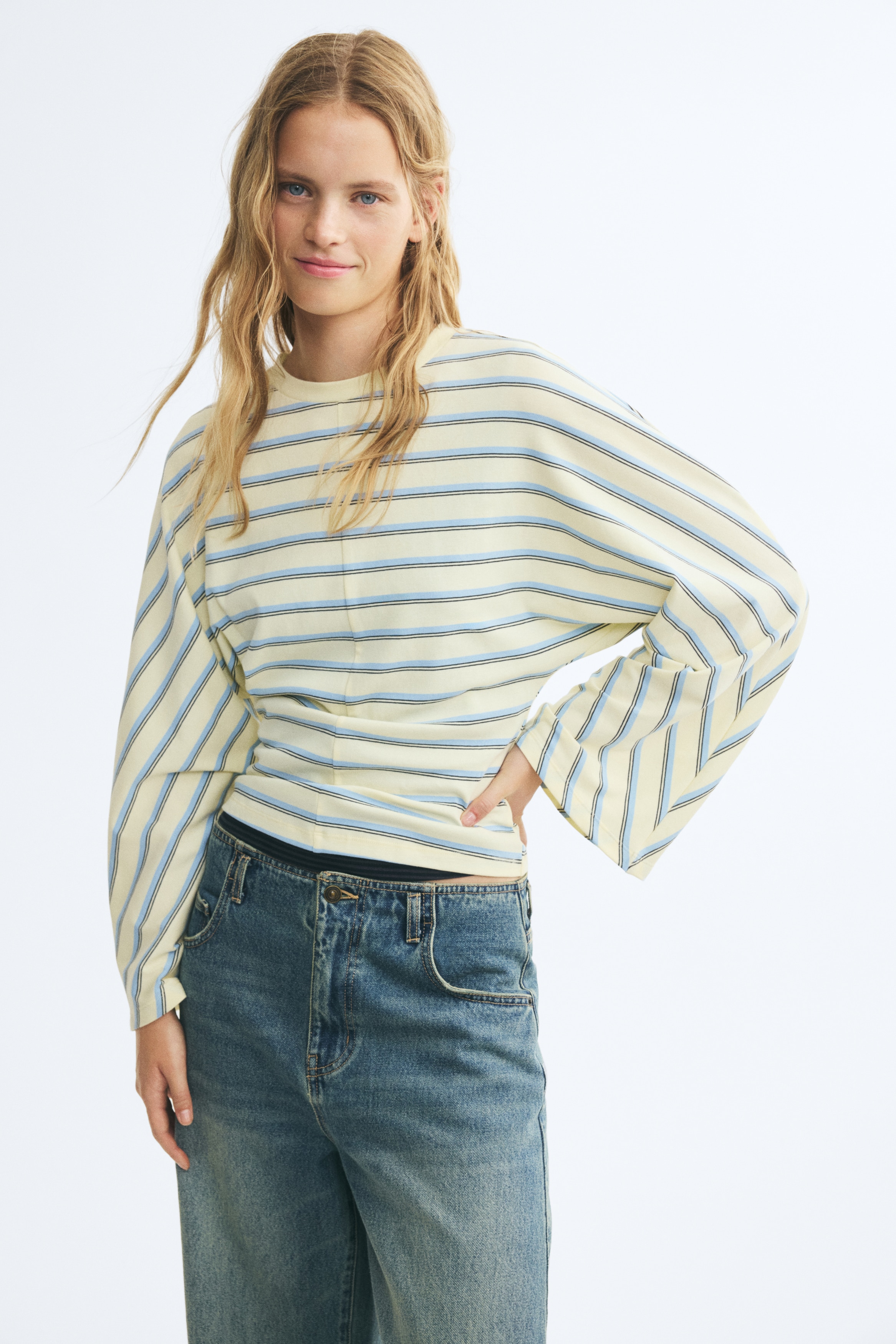 La parte superior de manga larga, vista de frente, presenta una silueta cropped en amarillo pálido, con un estampado de franjas horizontales azul claro y oscuras por el cuerpo y franjas diagonales en las mangas anchas y drapeadas. Incorpora un cuello redondo de canalé y una costura vertical que recorre el centro del delantero.