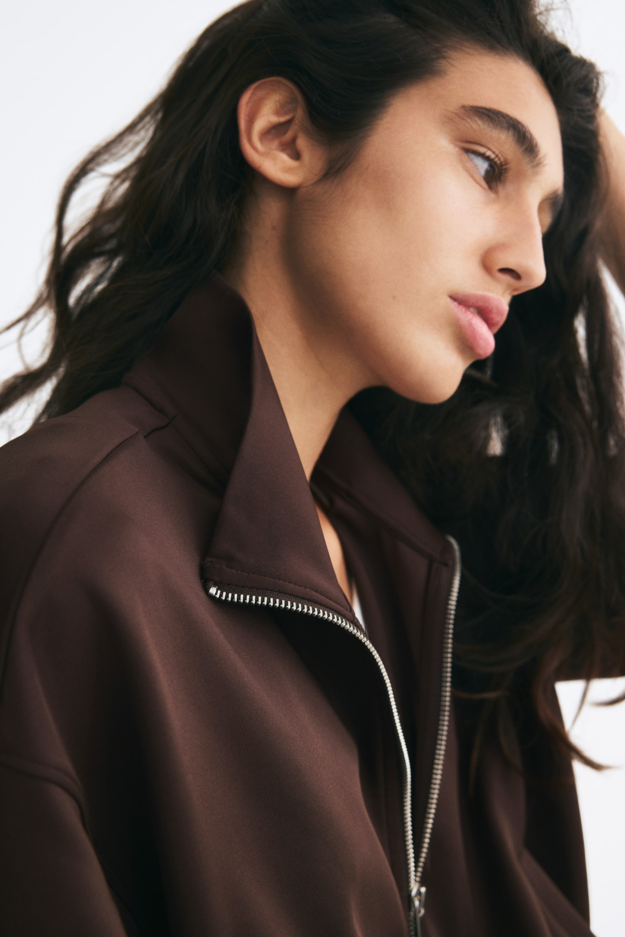 View larger image: The model is wearing a dark brown jacket in a fine ribbed jersey with a slight sheen, shown in a close-up profile view. The garment features a high stand collar, dropped shoulders, and a silver metallic front zipper that is partially open.