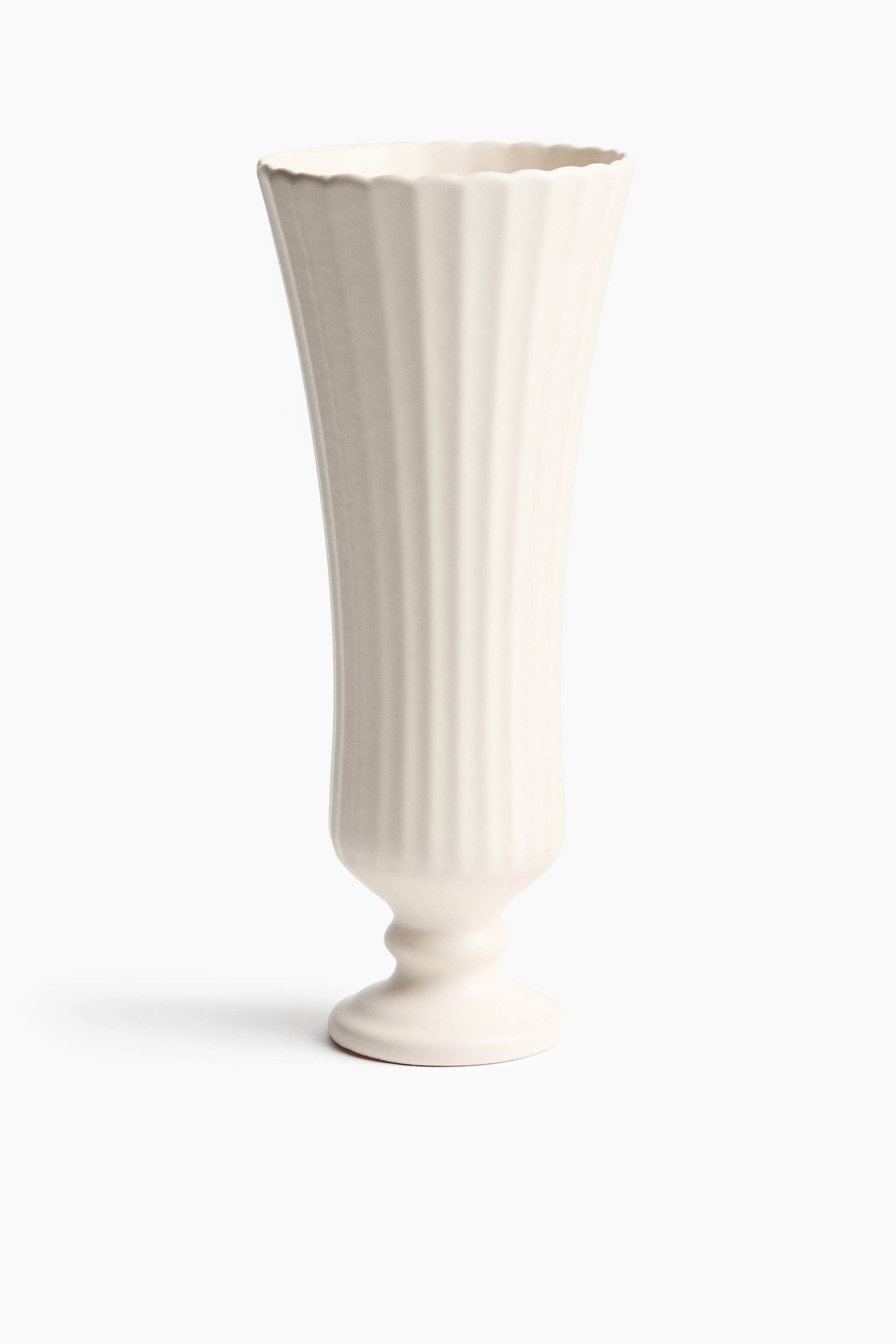 Agrandir l'image: Un vase en céramique de couleur crème est présenté de face, avec un corps haut et cannelé, orné de rainures verticales et d’un bord supérieur festonné. Le corps s’affine légèrement vers le bas avant de se prolonger en une base sur piédestal avec un pied circulaire évasé.