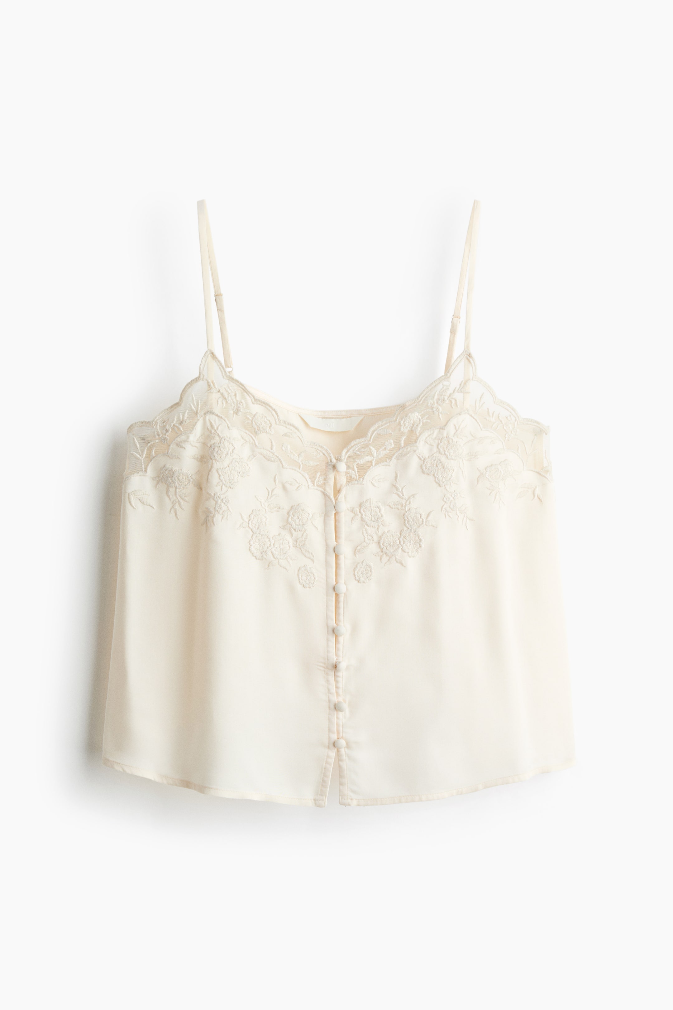 Grösseres Bild ansehen: Ein cremefarbener, gewebter Camisole wird in Frontansicht gezeigt und verfügt über einen V-Ausschnitt mit gewellter Spitzenborte und feiner Blumenstickerei. Das Oberteil zeigt schmale, verstellbare Träger sowie eine mittige Reihe kleiner, mit Stoff bezogener Knöpfe.