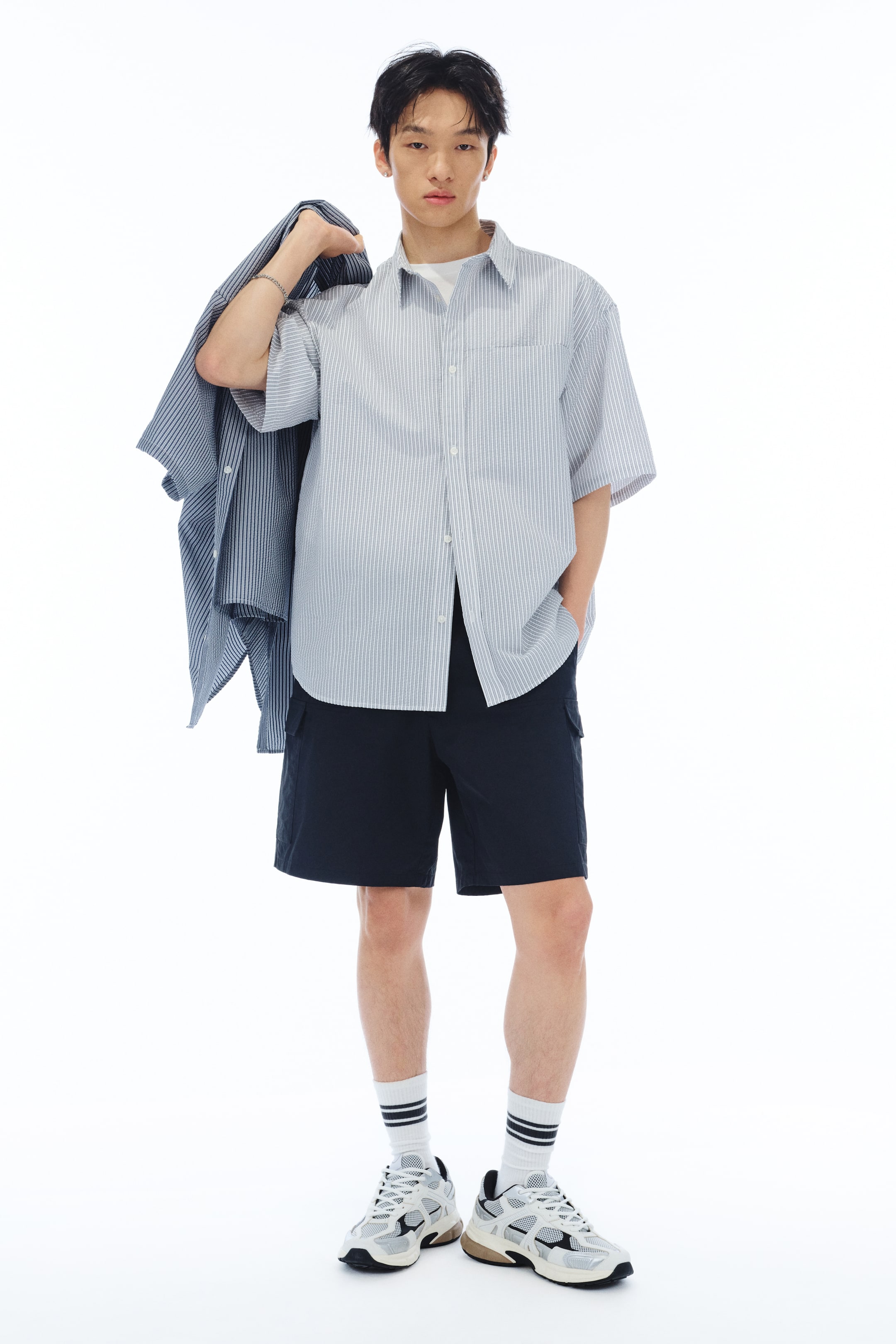 이미지 확대 보기: 모델은 흰색 크루넥 이너셔츠 위에 연한 파란색과 흰색 줄무늬의 반소매 버튼다운 셔츠를 착용하고 있습니다. 또한 모델은 왼쪽 어깨에 유사한 연한 파란색과 흰색 줄무늬의 긴소매 셔츠를 걸치고 있으며, 짙은 네이비색 카고 반바지를 입고 있습니다.