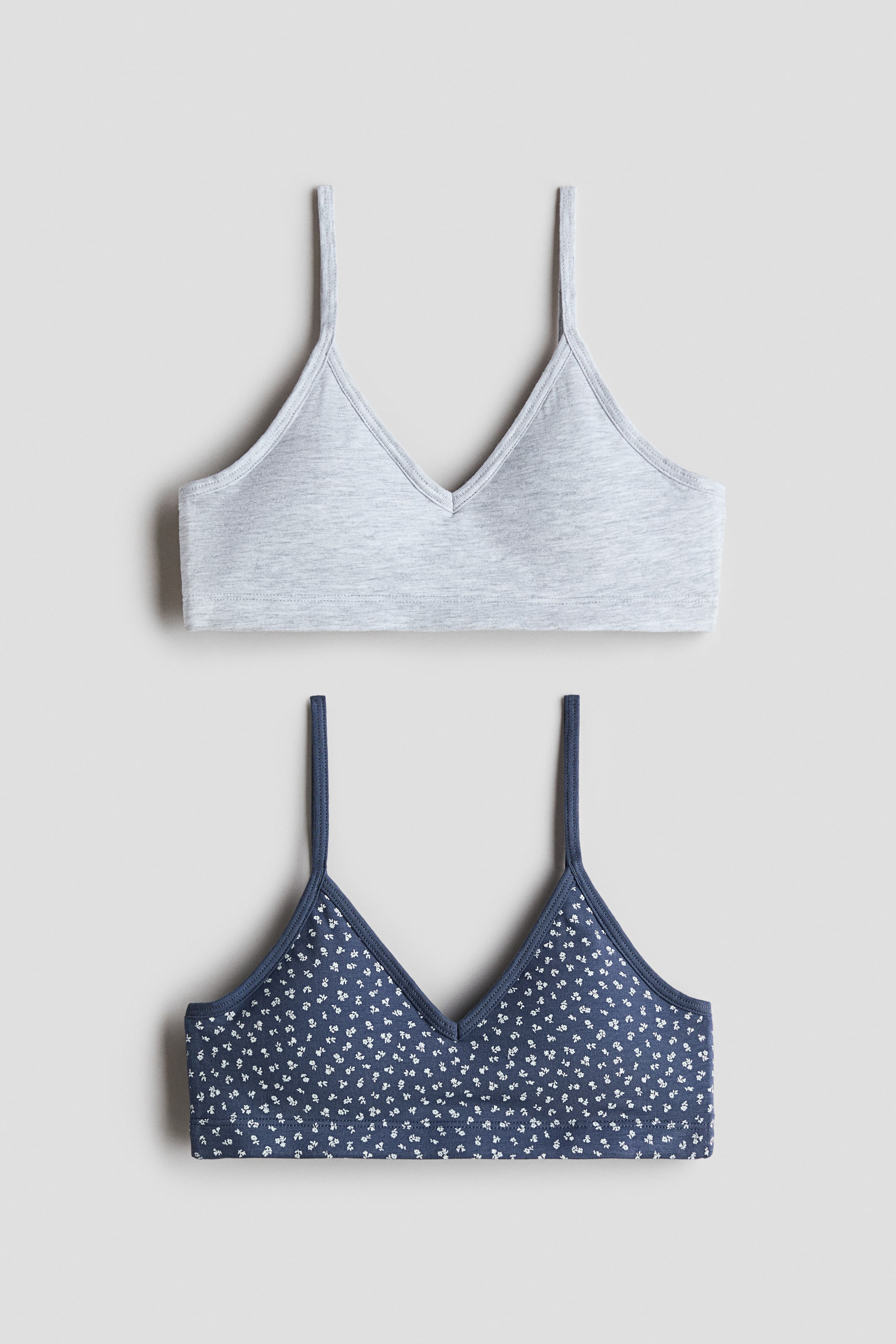 View larger image: Two bralettes are displayed flat, one above the other. The top bralette is heather grey, featuring a V-neck and spaghetti straps. The bottom bralette is dark blue with a small white floral print, also designed with a V-neck and spaghetti straps.