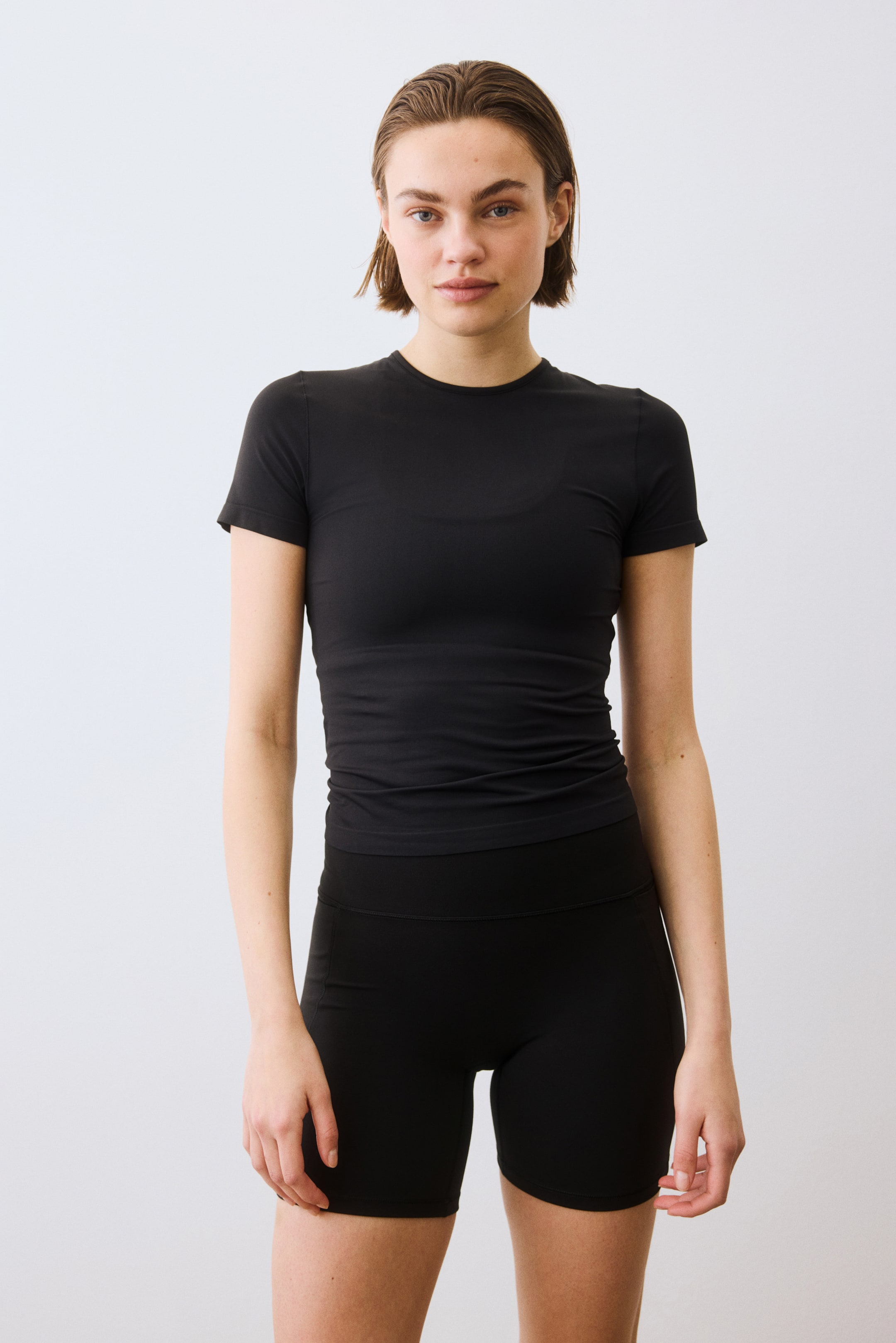 Ampliar la imagen: Una camiseta negra de cuello redondo y mangas cortas se ajusta ceñidamente al torso. Unos shorts negros a media pierna con una amplia pretina alta también se ajustan ceñidamente.