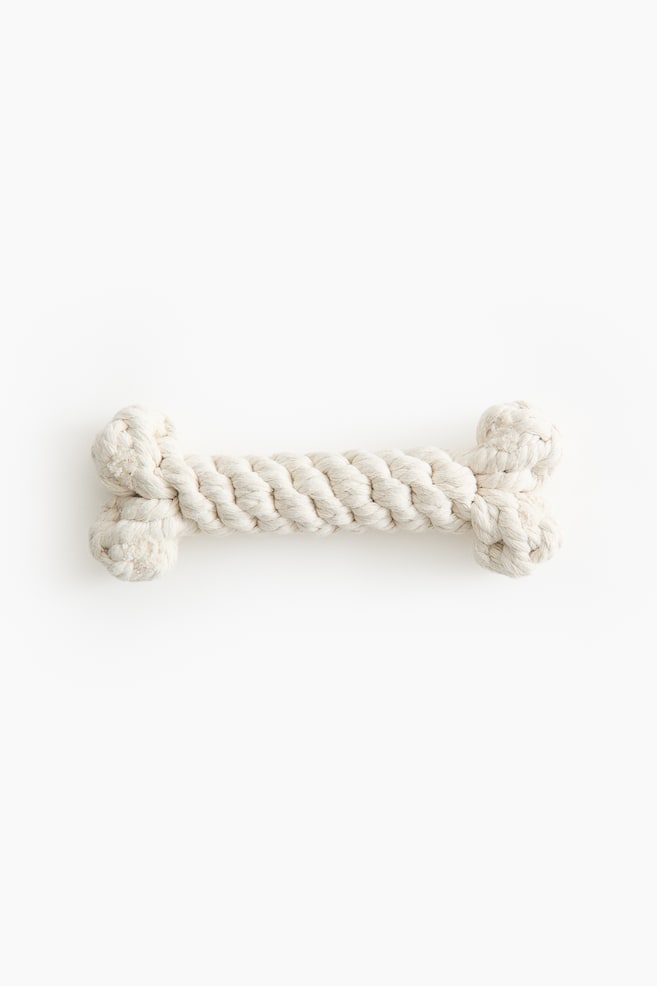 Un juguete para perro con forma de hueso, hecho de cuerda de algodón color blanquecino, está colocado de manera horizontal y orientado hacia el frente. El juguete muestra una barra central gruesa y trenzada, con extremos grandes y anudados que imitan las articulaciones de un hueso. - 1