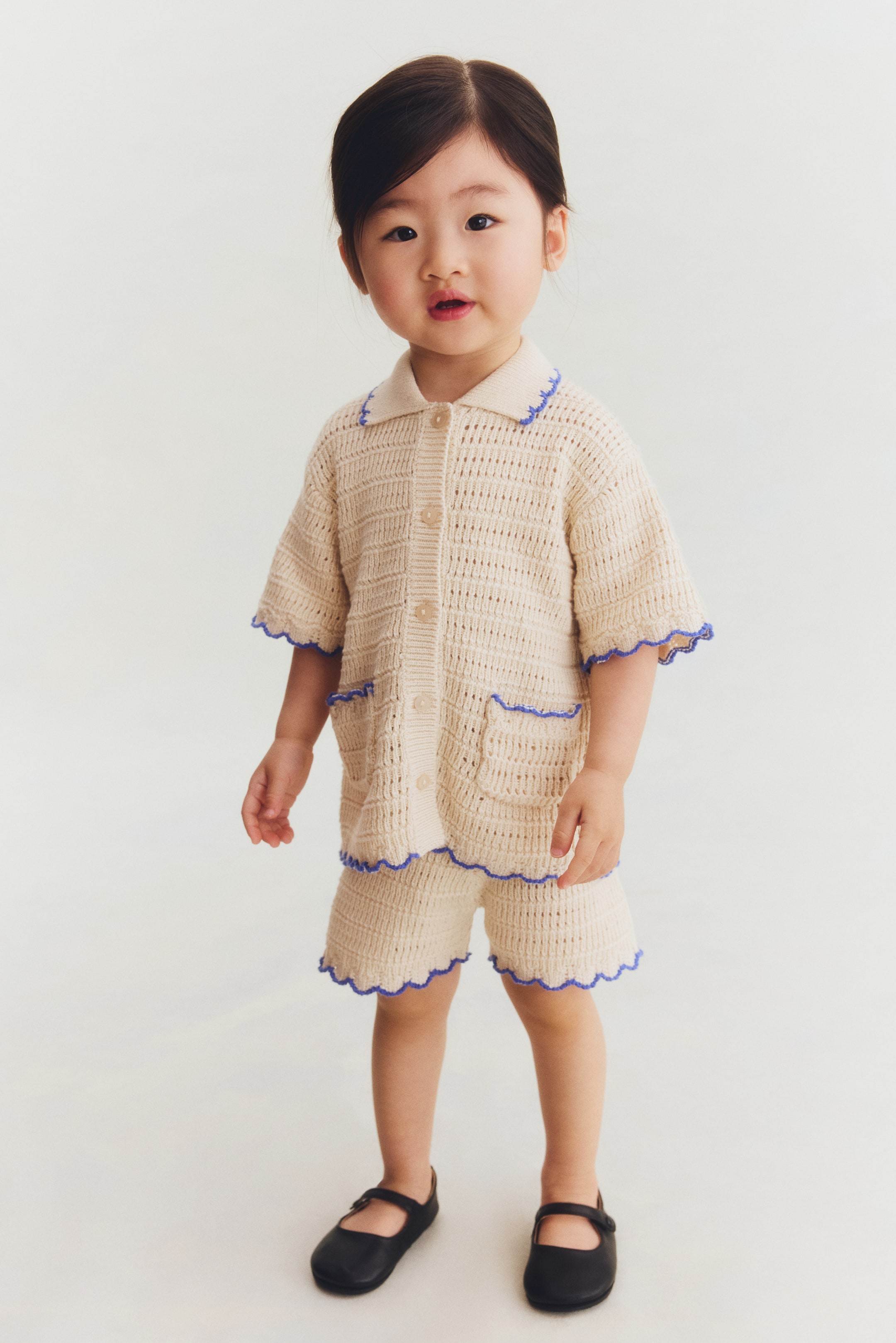View larger image: A cream open-knit two-piece set featuring a short-sleeve top and matching shorts, both detailed with scalloped blue trim. The top presents a pointed collar, a button-front closure, and two front patch pockets, while the shorts showcase scalloped leg openings.