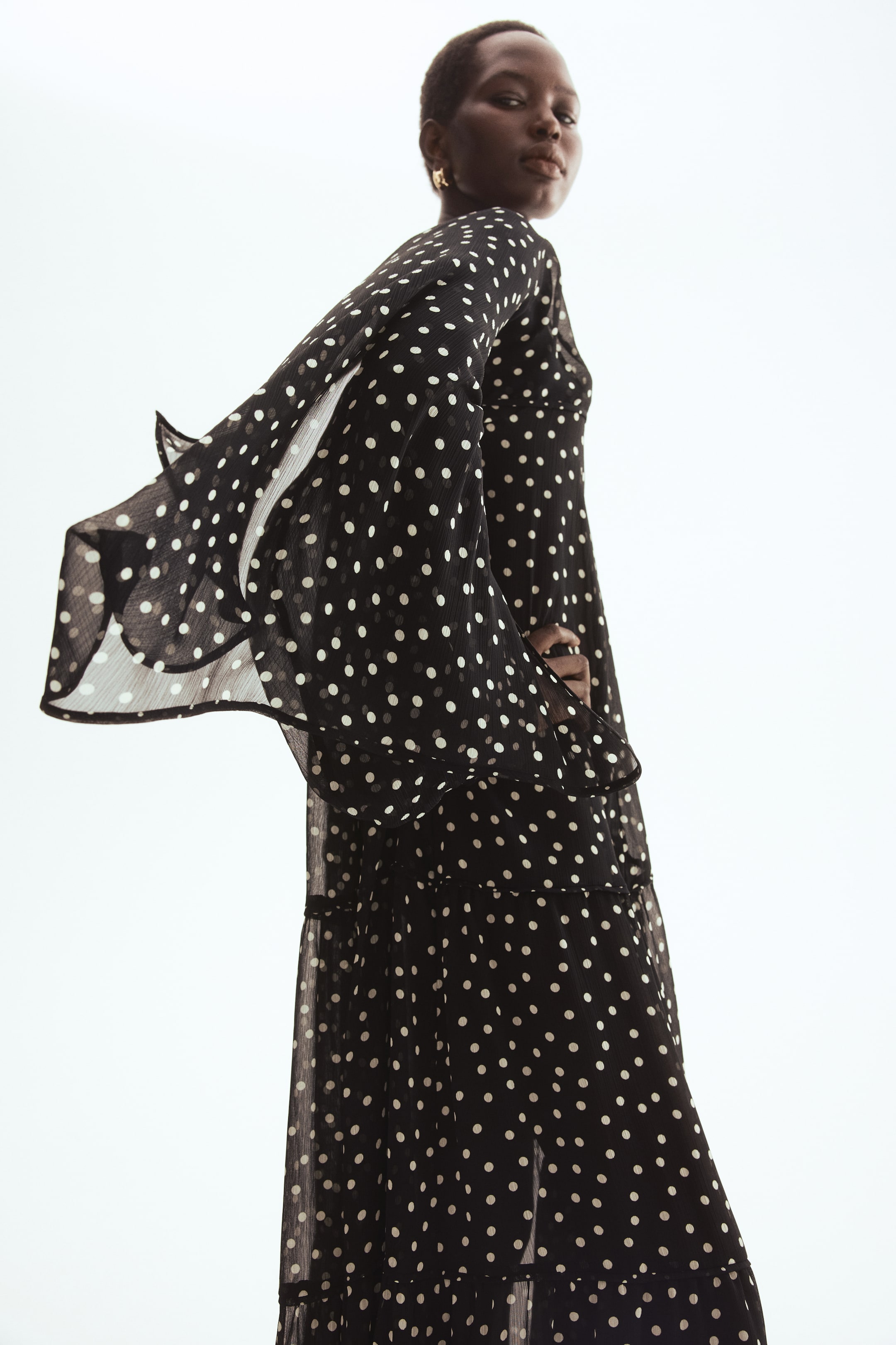 Grotere afbeelding bekijken: Een losvallende maxi-jurk in een doorschijnende zwarte stof met een witte polkadotprint wordt gedragen door het model. Het model poseert met één arm lichtjes omhoog, waardoor de lange, wijde mouwen en de asymmetrische cape van de jurk wapperen, terwijl geplooide lagen volume aan de rok toevoegen.