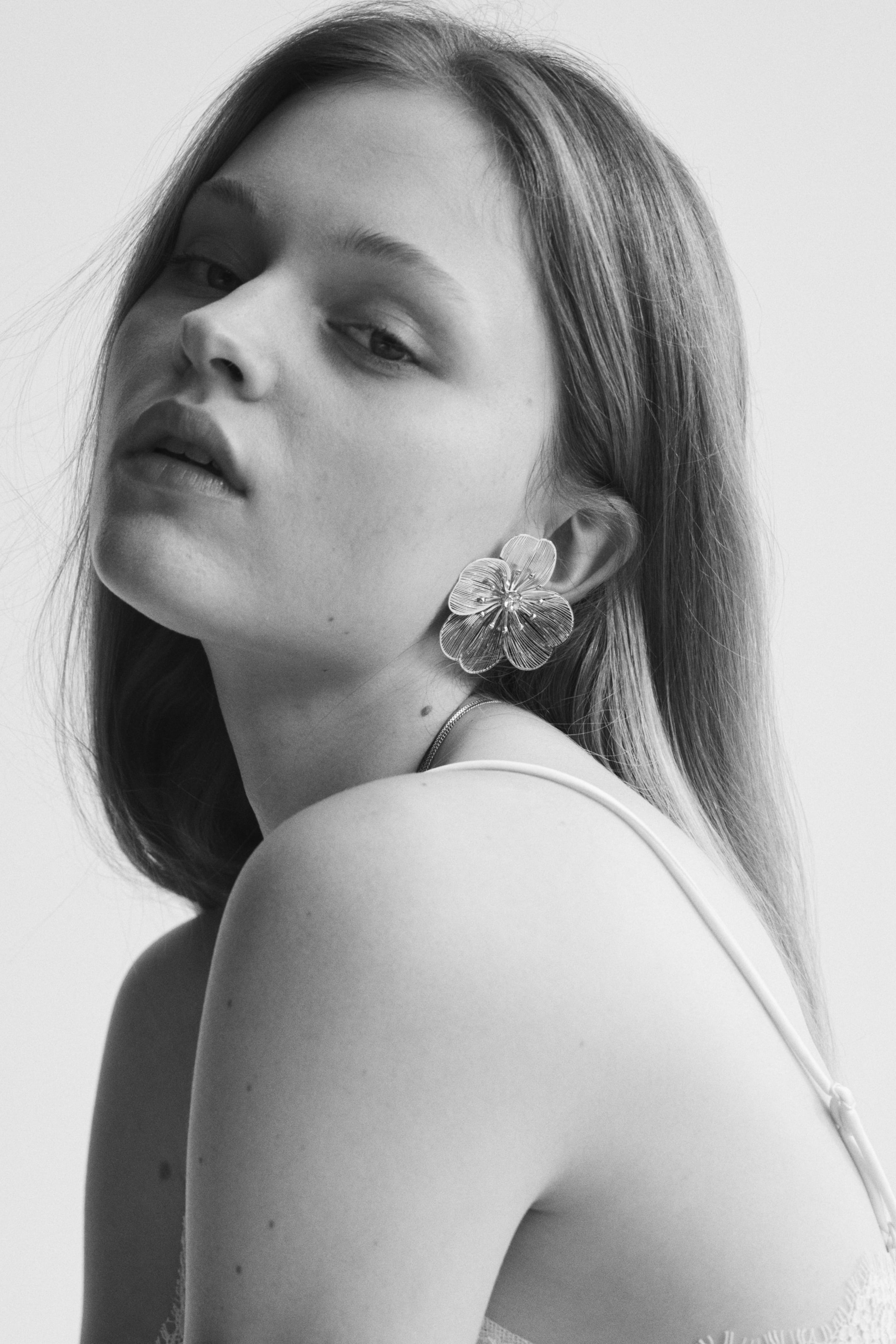 Zobacz większy obraz: Srebrzysty kolczyk w kształcie kwiatu zdobi ucho, lekko pochylony do przodu. Prezentuje wiele płatków wykonanych z delikatnych, drucikowych struktur oraz małe koralikowe detale w jego centrum.