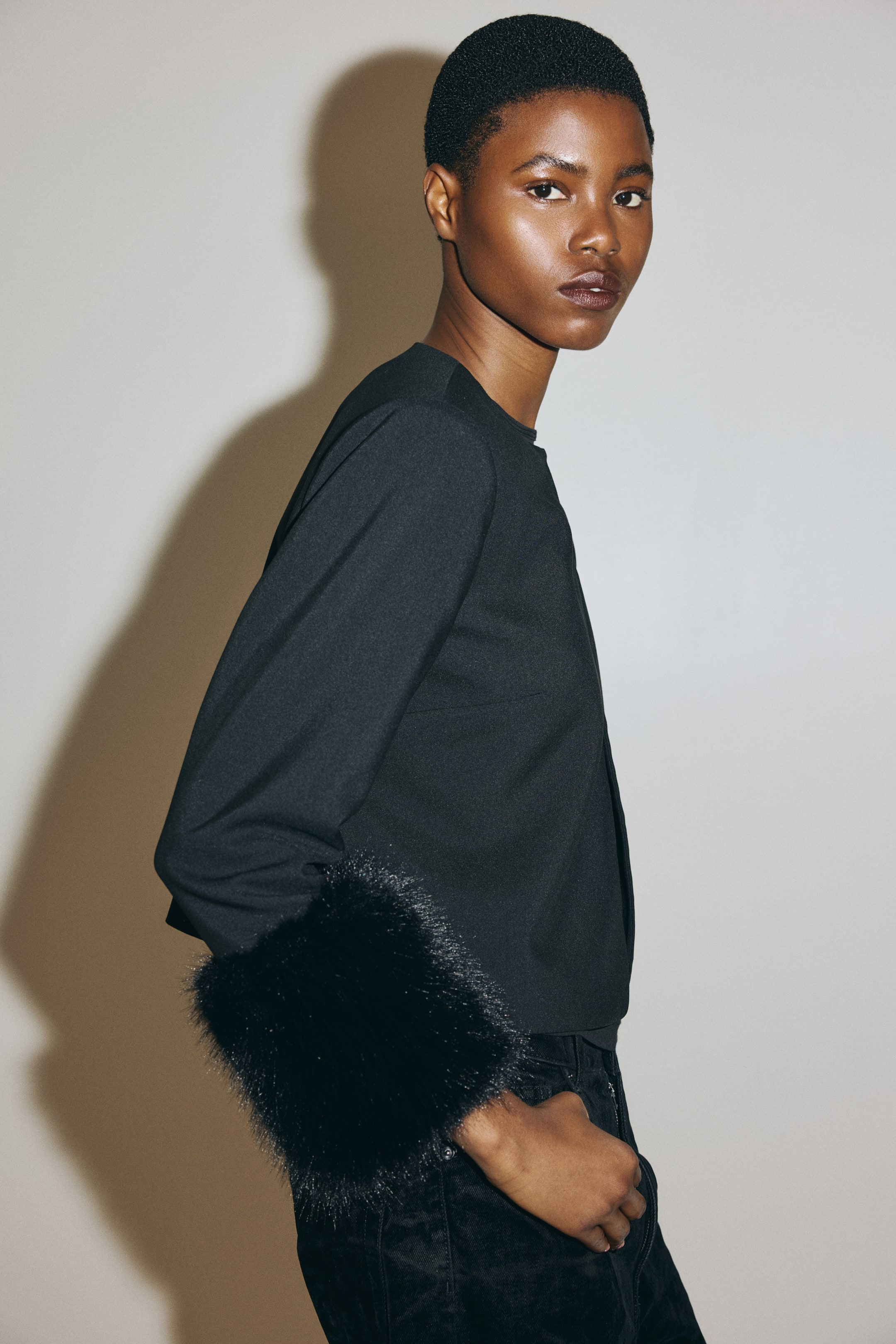 View larger image: A black long-sleeved top is being showcased from a side angle, featuring a sleek body and plush black faux fur cuffs.