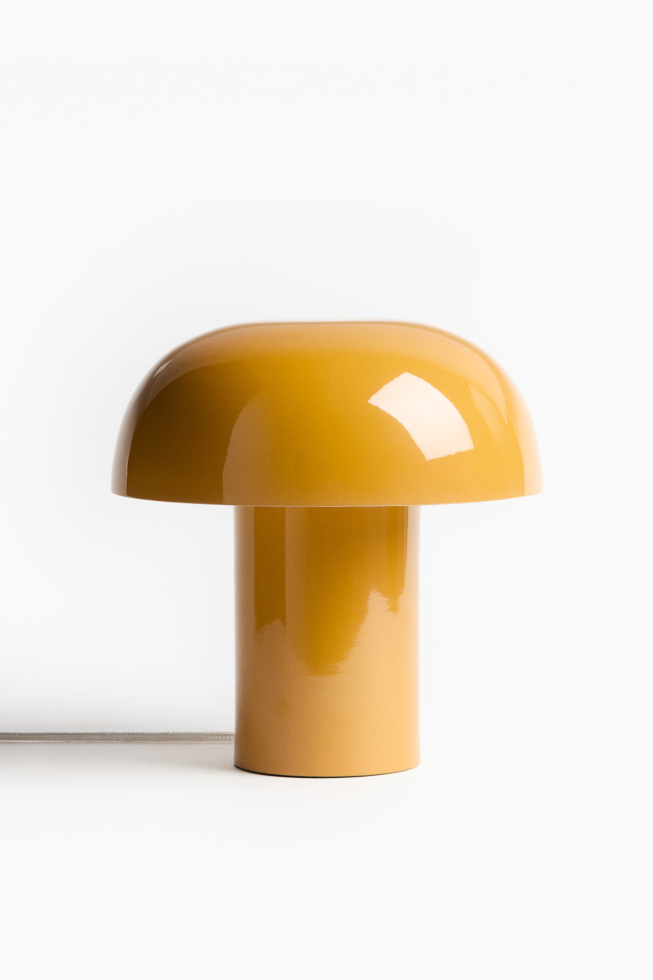 Visa större bild: En modern metallbordslampa presenterar en glansig, svampinspirerad design i senapsgul, med en rundad skärm som vilar på en cylindrisk bas, samt en tygklädd sladd som kommer ut från dess nedre sida.