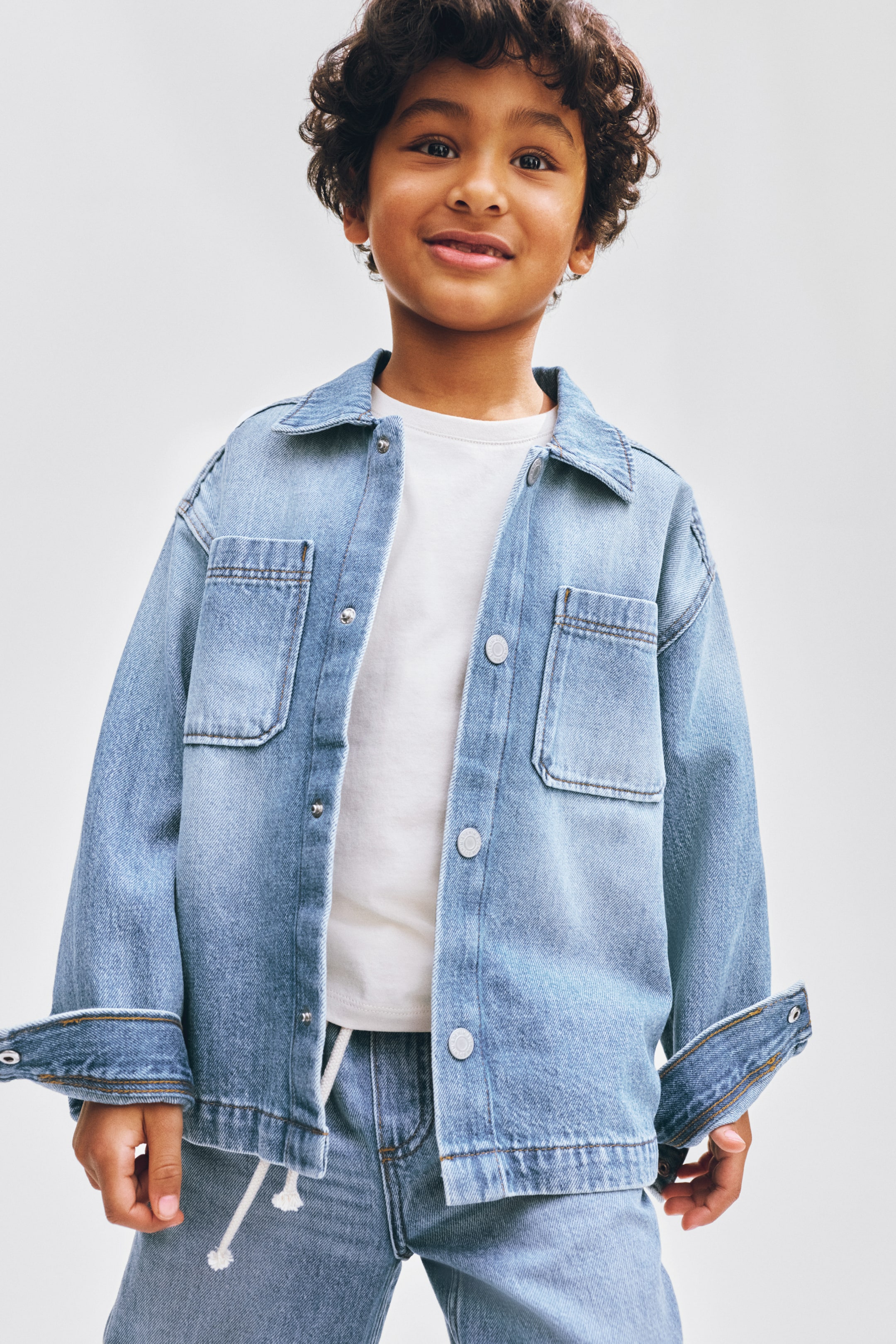 View larger image: A light blue denim jacket featuring a pointed collar, a button-up front with visible snaps, and two chest pockets with buttoned flaps, is layered over a white crew neck t-shirt and paired with light blue denim pants featuring a white drawstring.