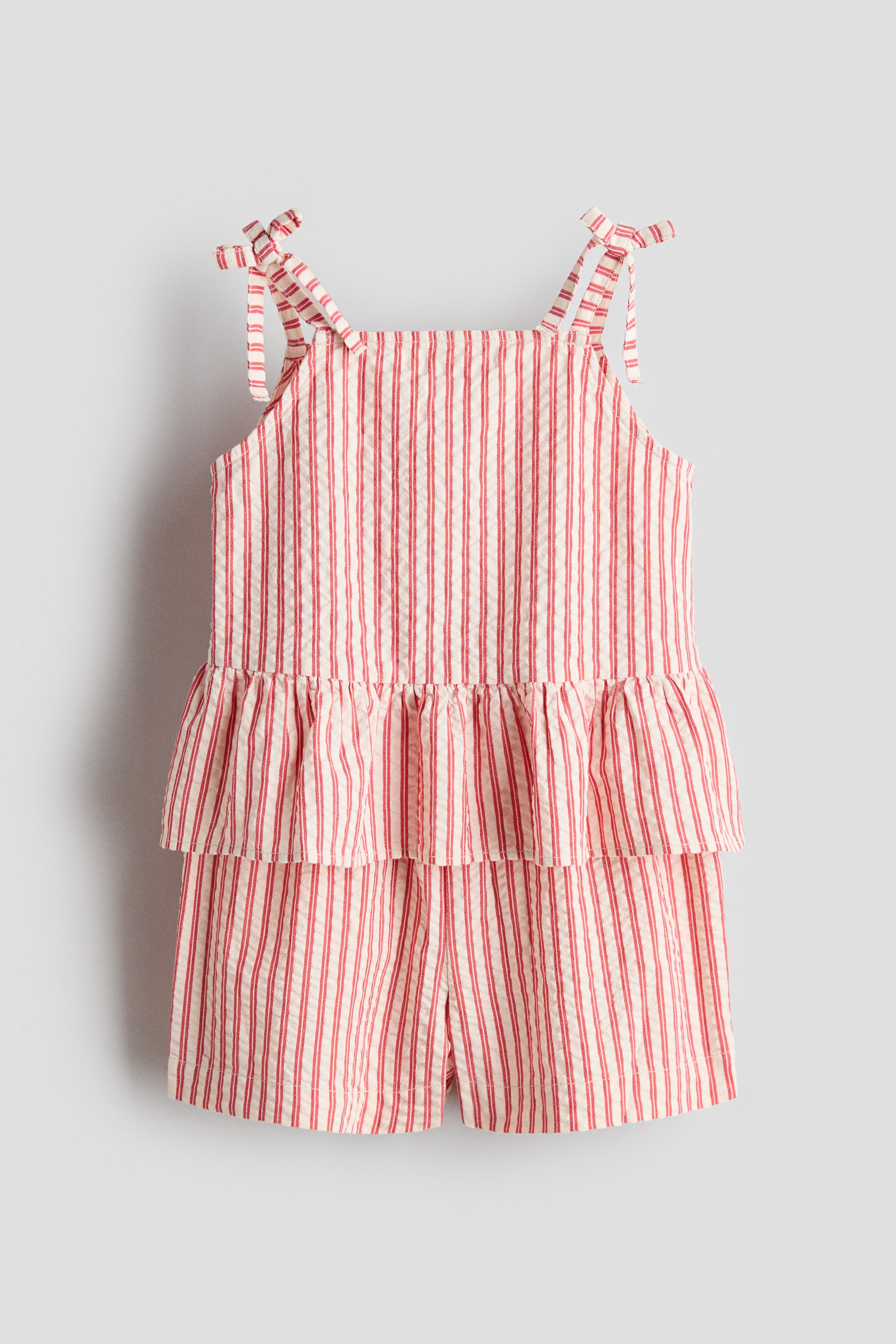 View larger image: A set shown from the front features a red-and-white vertically striped top and matching shorts in a textured cotton weave. The top is designed with a square neckline, narrow shoulder straps tied into decorative bows, a gathered waist and a flared peplum, while the shorts have an elasticated waist.