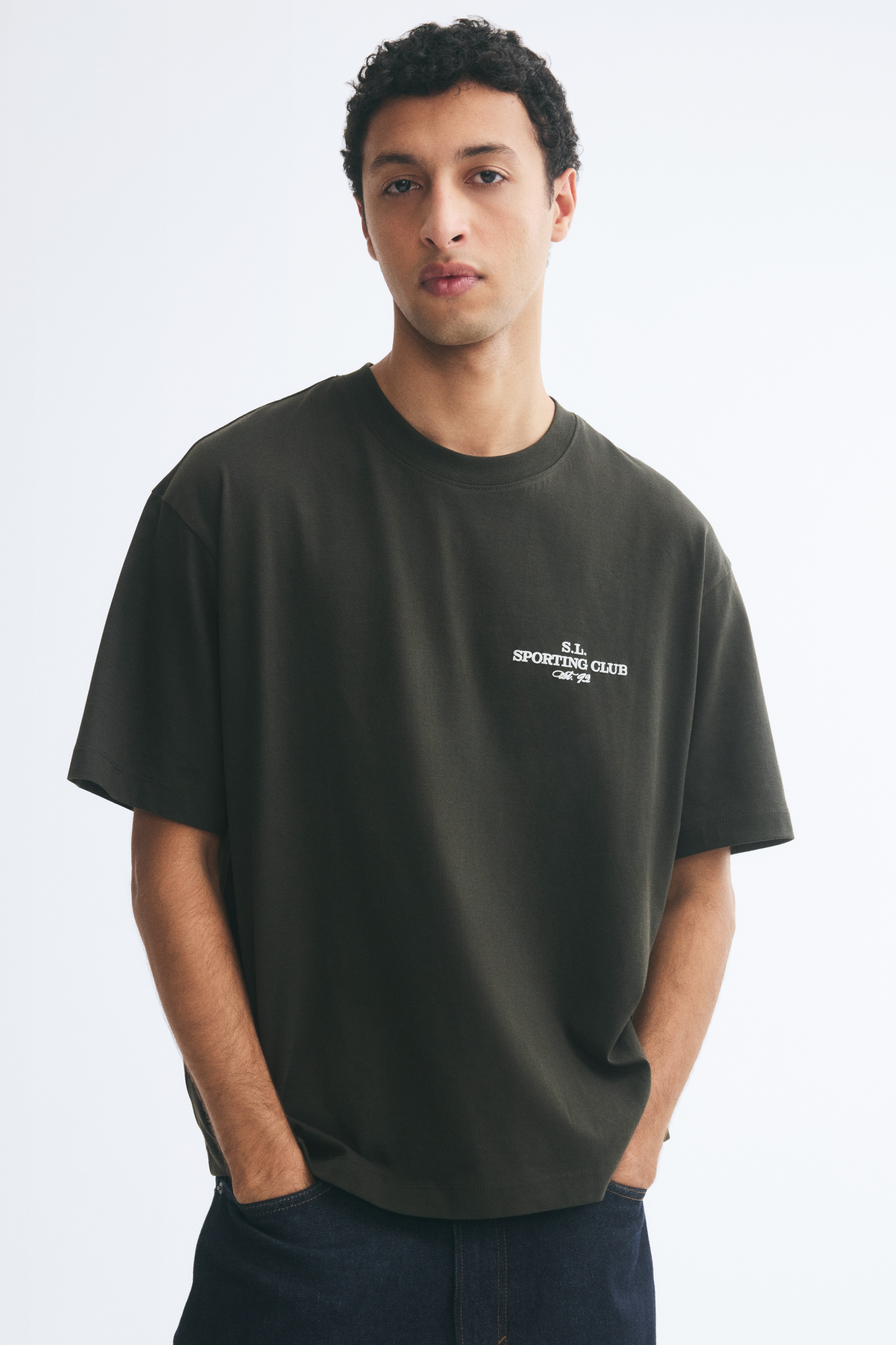 Má»t chiếc áo thun cá» tròn oversized mà u xanh lá Äáºm, ÄÆ°á»£c chụp chÃnh diá»n, vá»i tay ngắn rá»ng và gấu áo thẳng. á» ngá»±c trái có in dòng chữ Äá» há»a mà u trắng nhá» ghi âS.L. SPORTING CLUBâ.