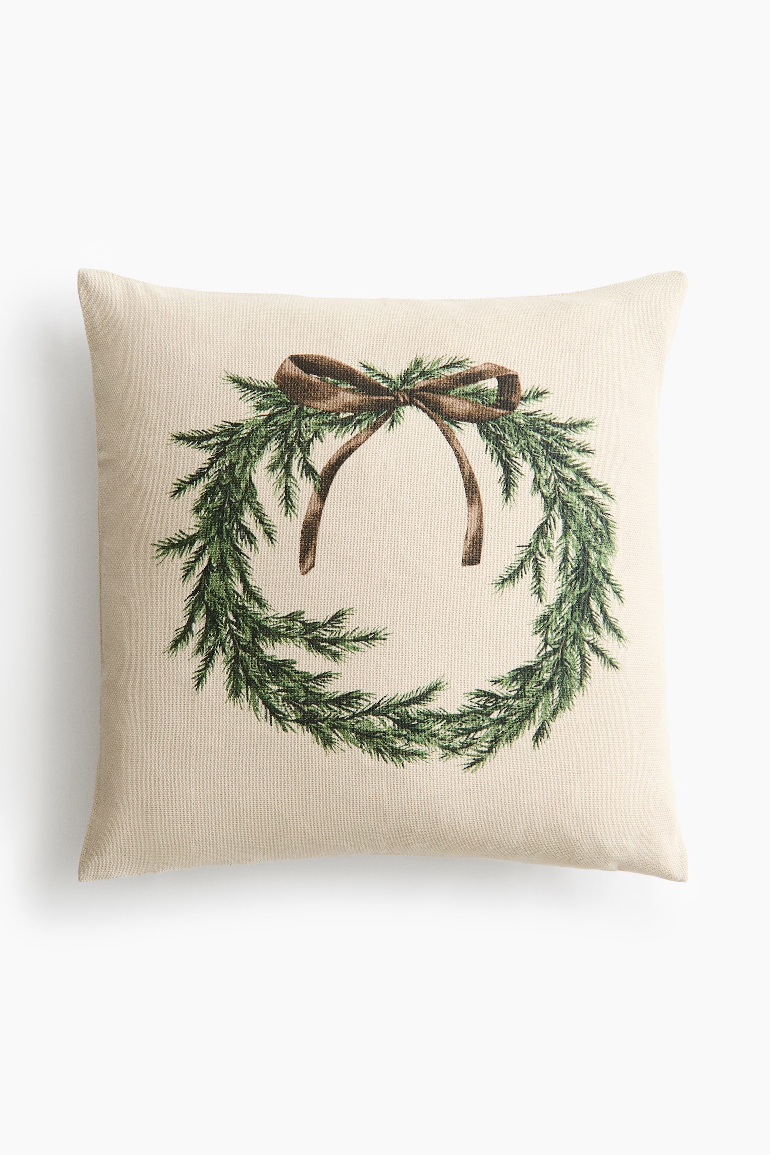 A square, cream-coloured throw pillow is facing the front, showcasing a printed design of a green pine branch wreath tied with a brown ribbon bow at the top. The pillow is made of a textured, canvas-like fabric.