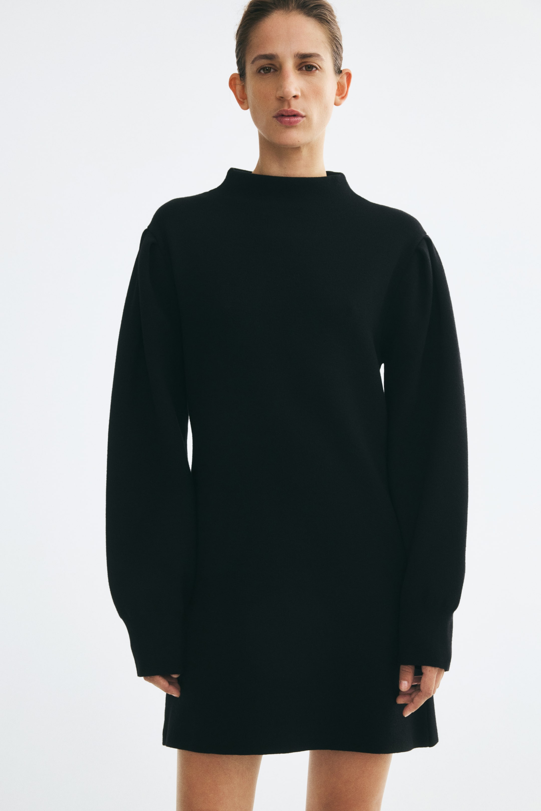 Voir une image plus grande: Une mini-robe noire en tricot côtelé avec un col montant et de longues manches bouffantes est portée par le mannequin.
