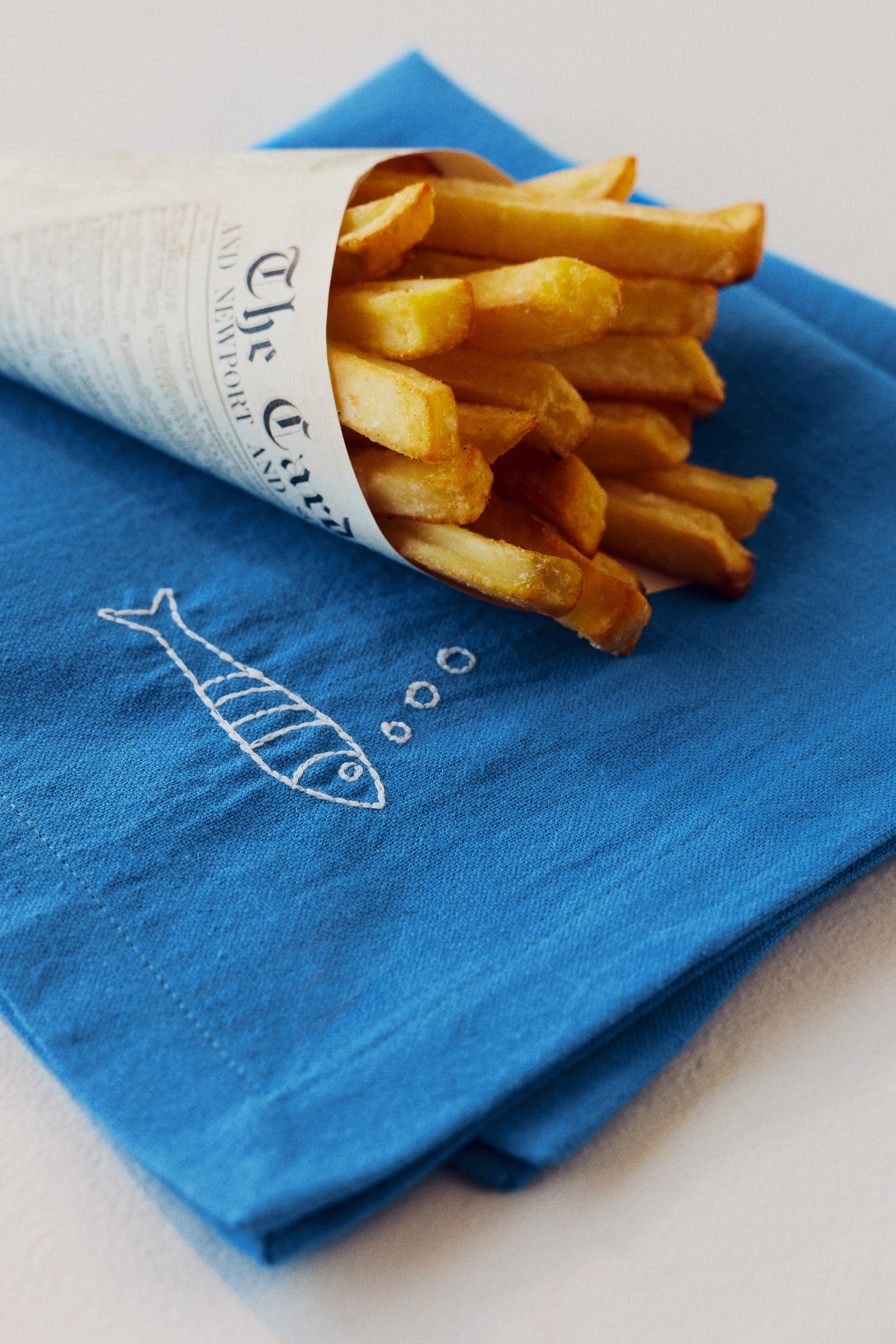 Grotere afbeelding bekijken: Goudbruine frieten vullen een kegelvormige verpakking gemaakt van papier dat lijkt op krantenprint, waarbij enkele frieten over de rand steken. De verpakking rust op een blauwe stof met een wit geborduurd visontwerp met bubbels.