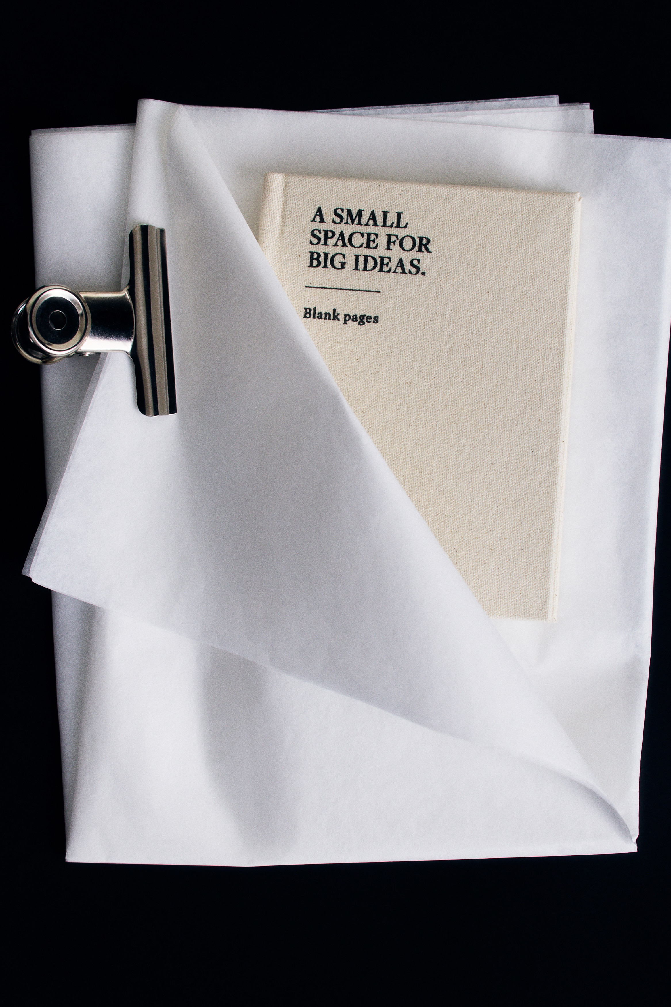 A small notebook featuring an off-white cotton cover is lying flat in a front view, showcasing the black printed text âA SMALL SPACE FOR BIG IDEAS.â and âBlank pages.â A thick black elastic band runs vertically along the right side to secure the cover.