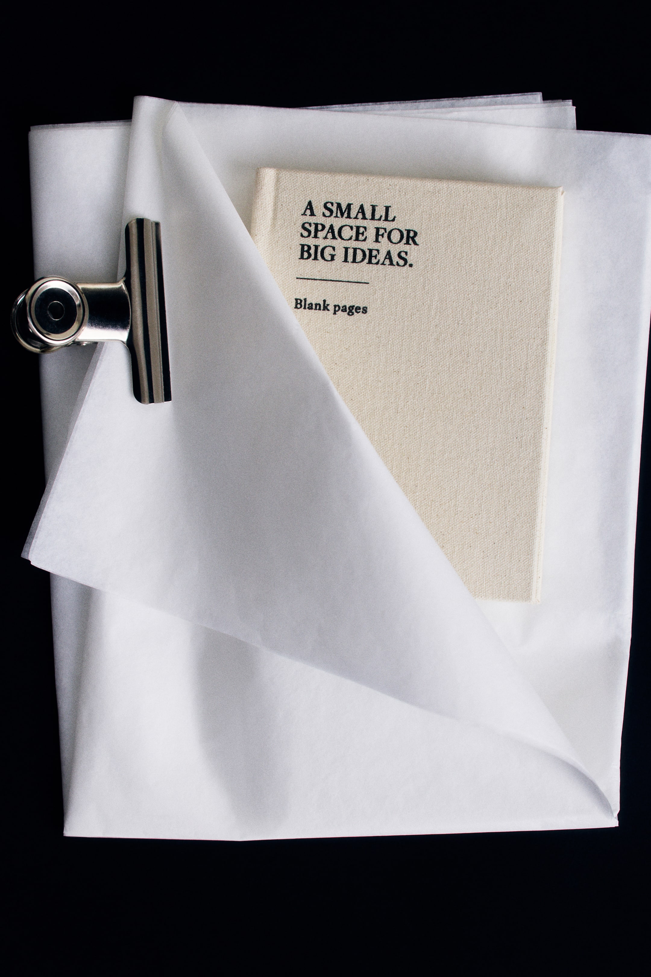 View larger image: A small notebook featuring a textured, cream-colored cotton cover lies flat, partially wrapped in folded white tissue paper secured on the left with a silver metal binder clip. The notebook’s front cover displays the printed black text, “A Small Space for Big Ideas.” and “Blank pages.”