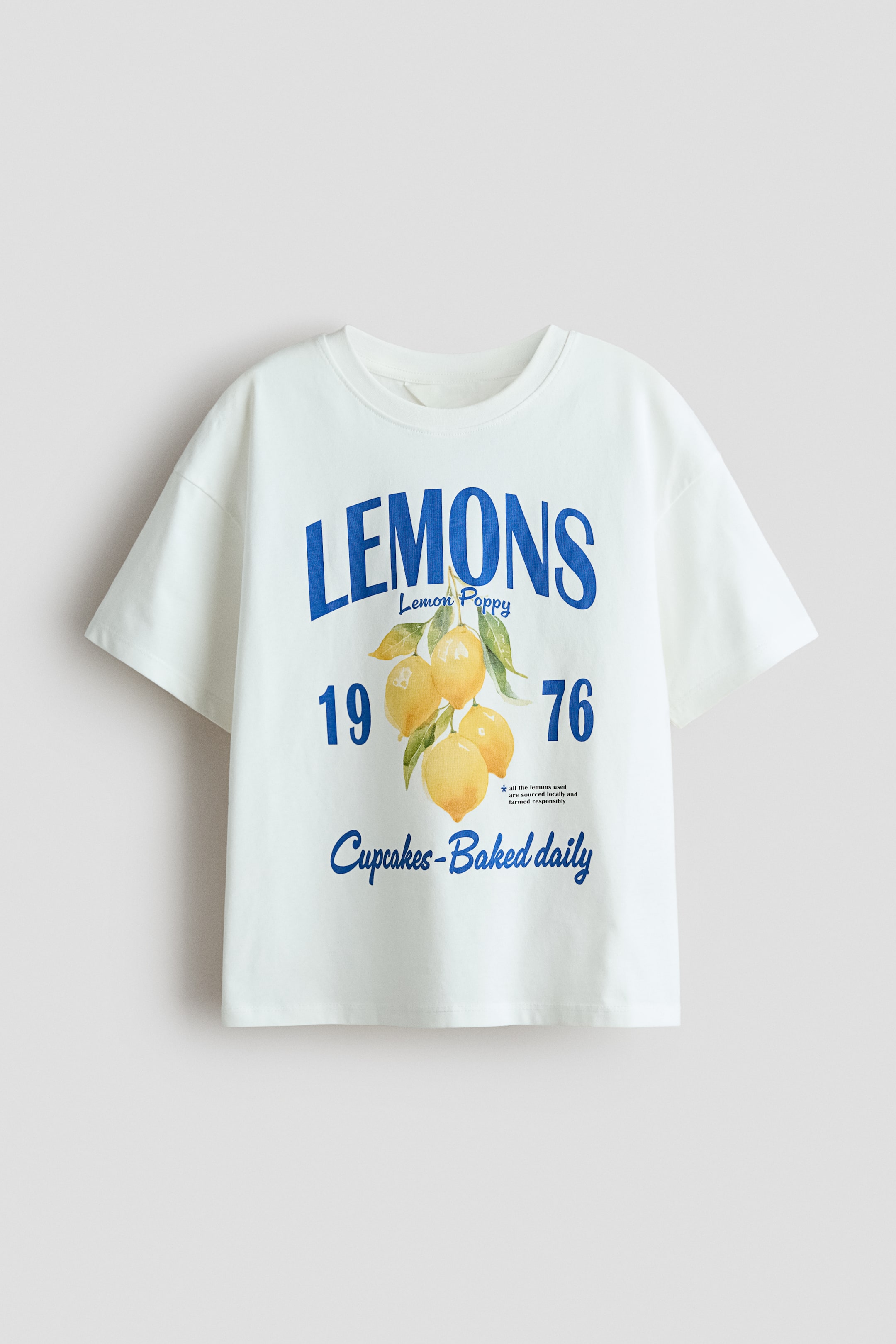 View larger image: An oversized t-shirt in soft, printed cotton jersey is shown from the front, featuring a round, ribbed neckline, dropped shoulders, and a straight-cut hem. The front displays a graphic with "LEMONS" in large blue letters, "Lemon Poppy" in smaller blue text, an illustration of yellow lemons, the blue numbers "19" and "76", and "Cupcakes-Baked daily" in blue cursive text.