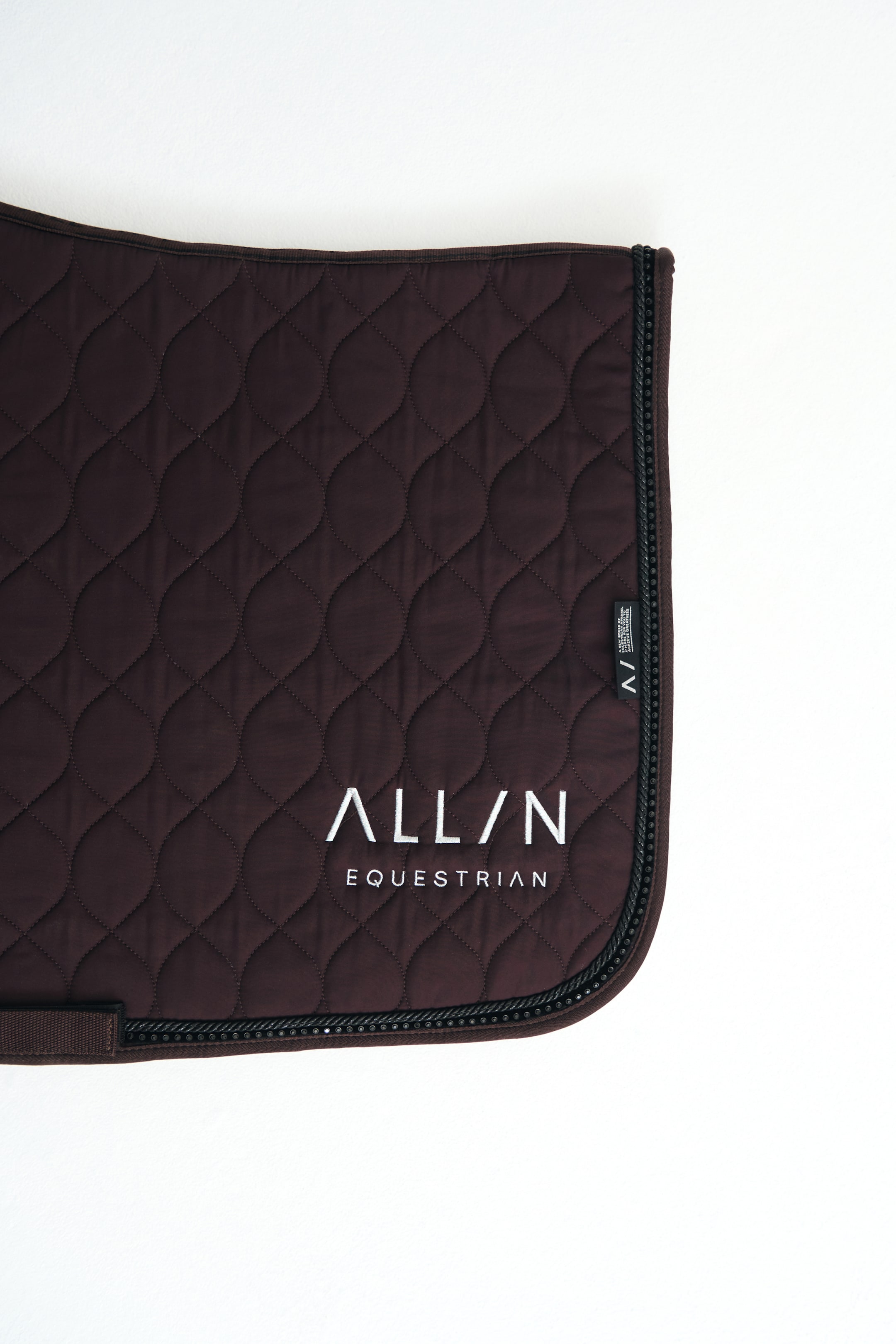 View larger image: A dark brown quilted saddle pad features an ogee-style quilted pattern and a white embroidered logo reading "ALL/N EQUESTRIAN" near the bottom corner. The pad showcases a rounded edge detailed with black braided cord piping and a sparkling rhinestone trim.