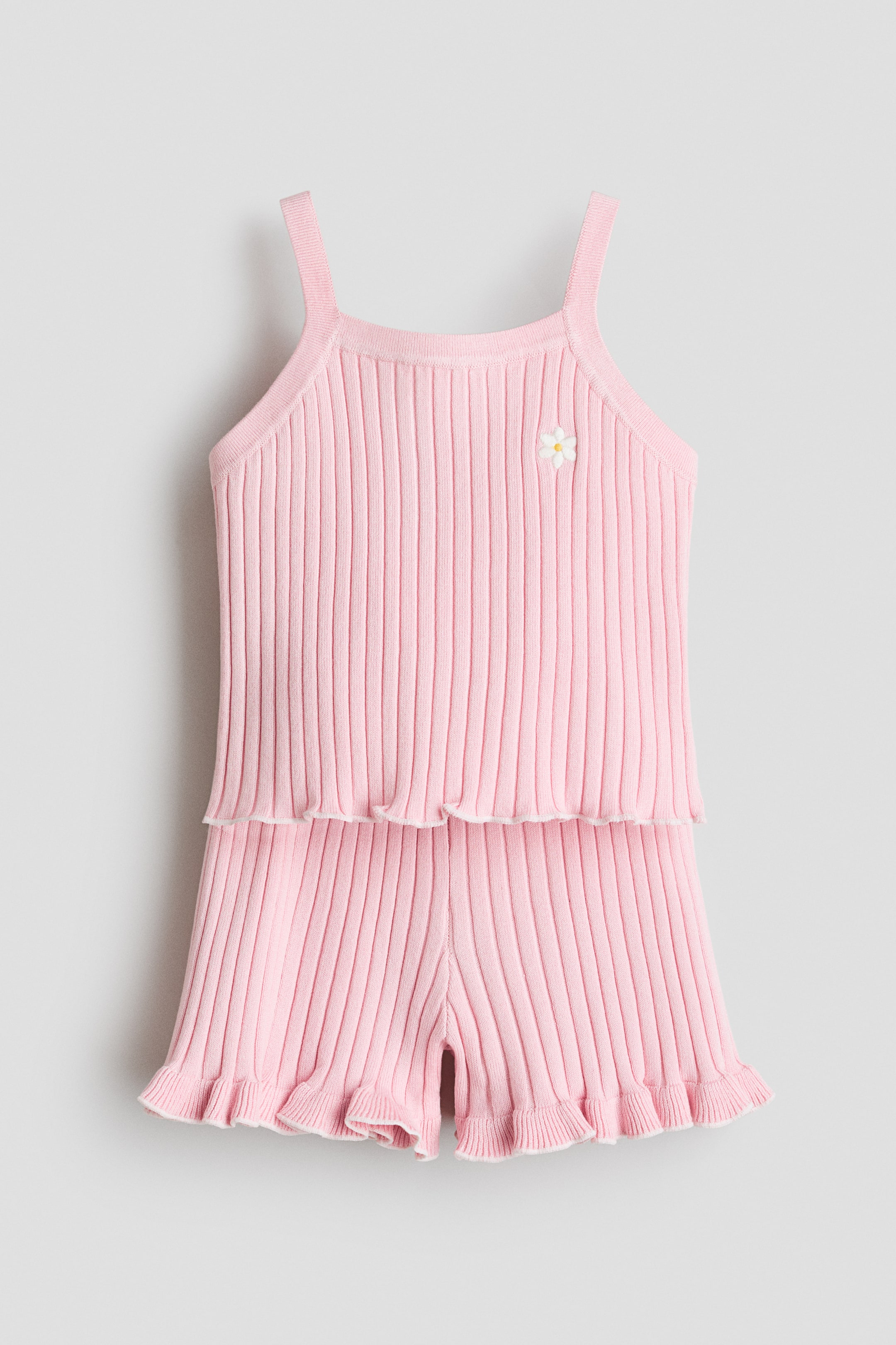 View larger image: A pink ribbed knit set is showcased from the front, featuring a strappy top with a square neckline and a small white flower appliqué on the chest. The top's bottom edge is overlocked, complementing the frill-trimmed hems of the shorts.