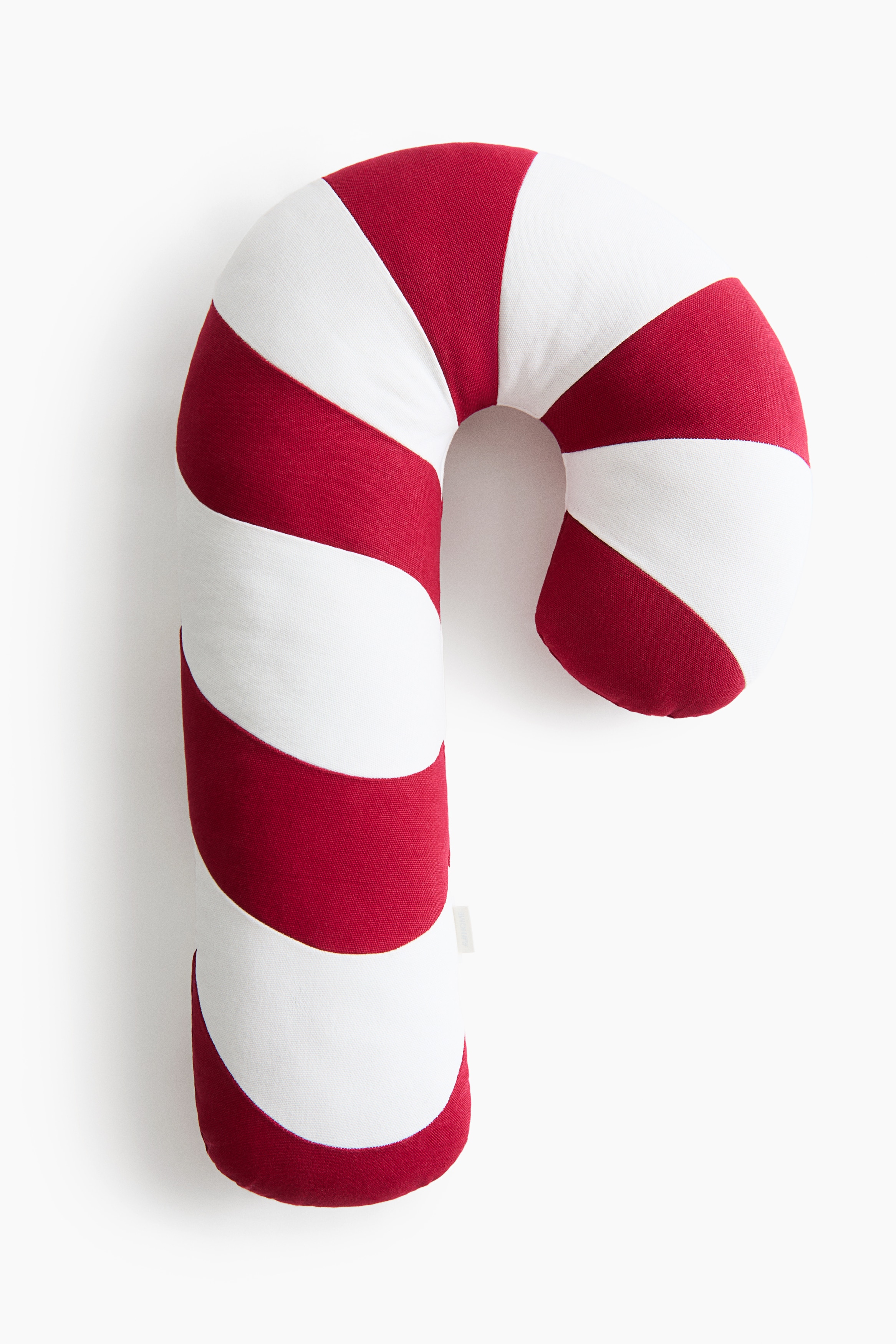 A candy cane-shaped cushion in cotton canvas is resting upright on a bed, showcasing wide, alternating red and off-white stripes. The plush, curved cushion is leaning against a pillow, displaying its rounded ends and textured surface.