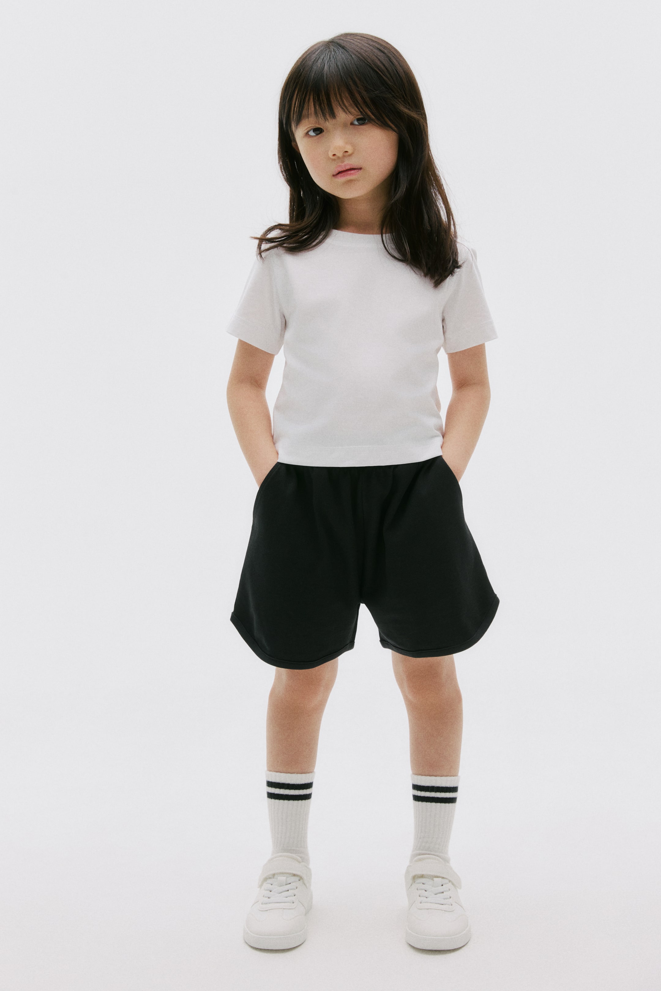 View larger image: A white short-sleeved t-shirt and black athletic shorts are being worn by the model, who is placing hands into the shorts' pockets. White crew socks with two black stripes are complementing white low-top sneakers with hook-and-loop closures.