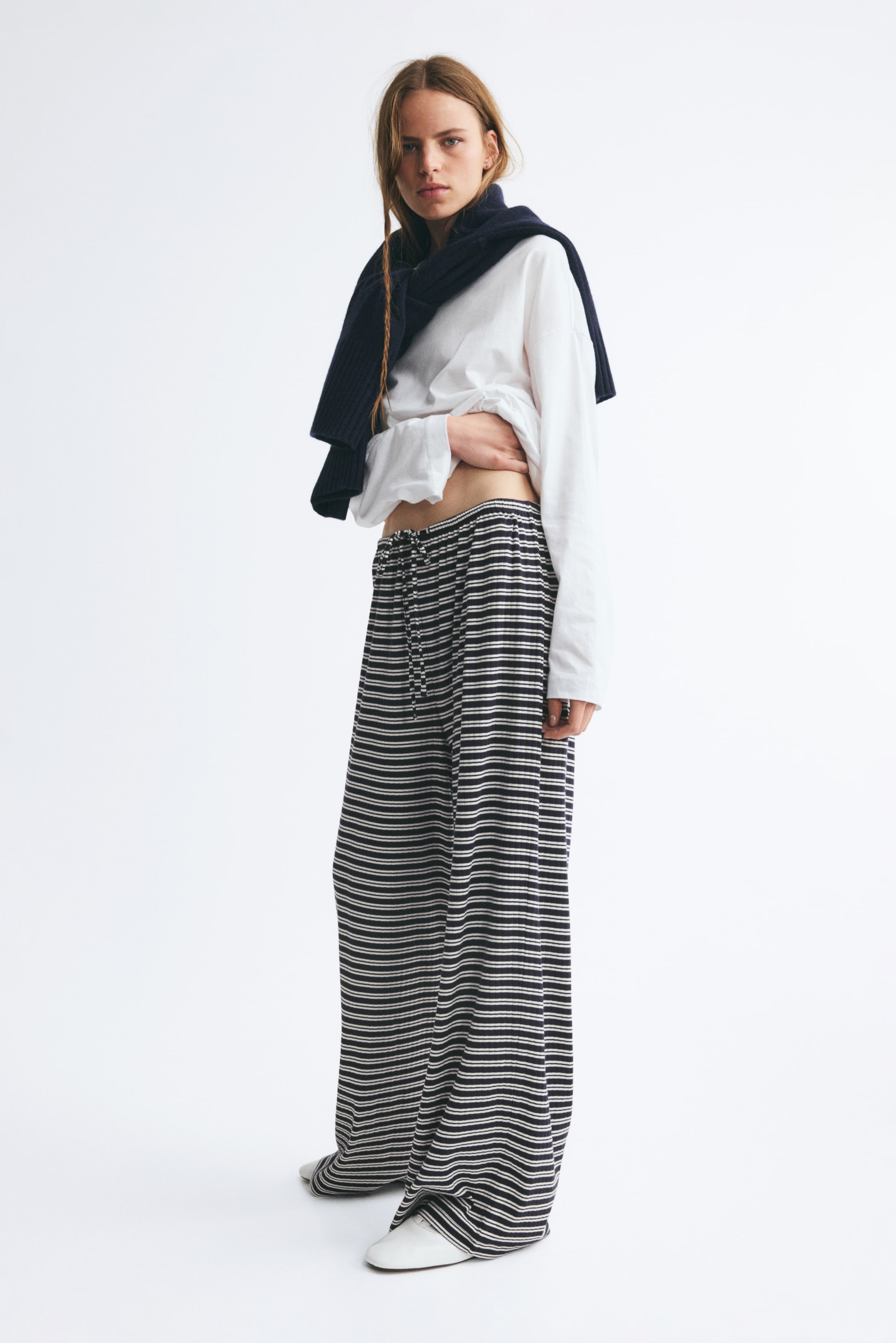 View larger image: Striped, loose-fit trousers in ribbed cotton jersey are being worn by the model. The model is lifting the hem of a white long-sleeved top to reveal the elasticised, drawstring waistband of the trousers.