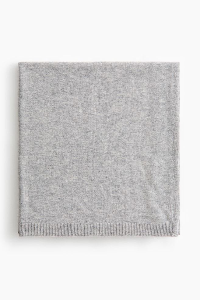 A baby blanket in soft, light gray fine knit wool is lying folded flat in a square shape, showcasing its front view. The blanket is featuring a ribbed knit trim along the bottom edge. - 1