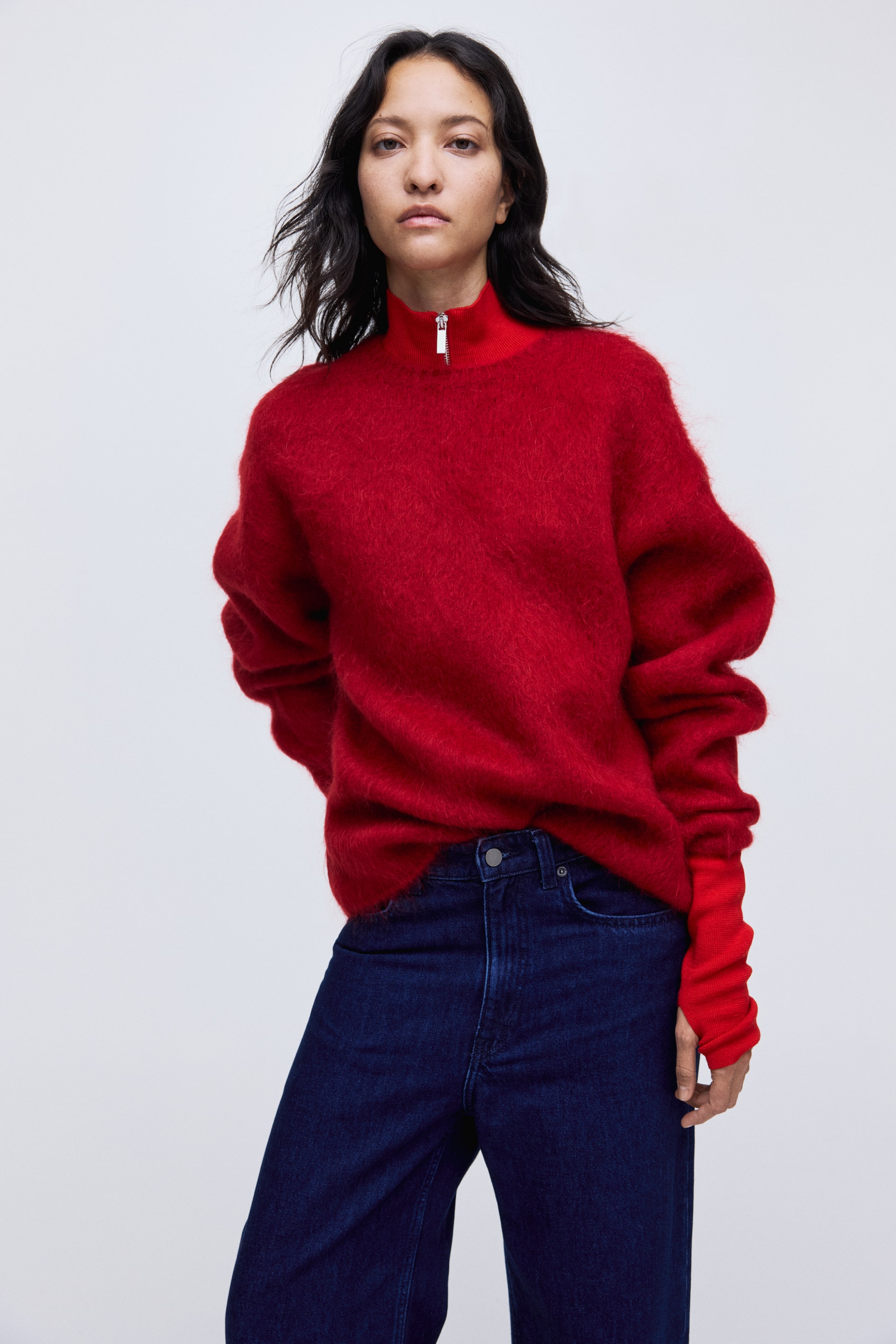 Ampliar la imagen: Un suéter de punto cepillado rojo se presenta desde el frente, ligeramente inclinado hacia la derecha, y cuenta con un cuello alto acanalado que se abre con un cierre de media cremallera en tono plateado. Sus mangas largas y sueltas se extienden hacia puños acanalados, cubriendo parcialmente las manos.