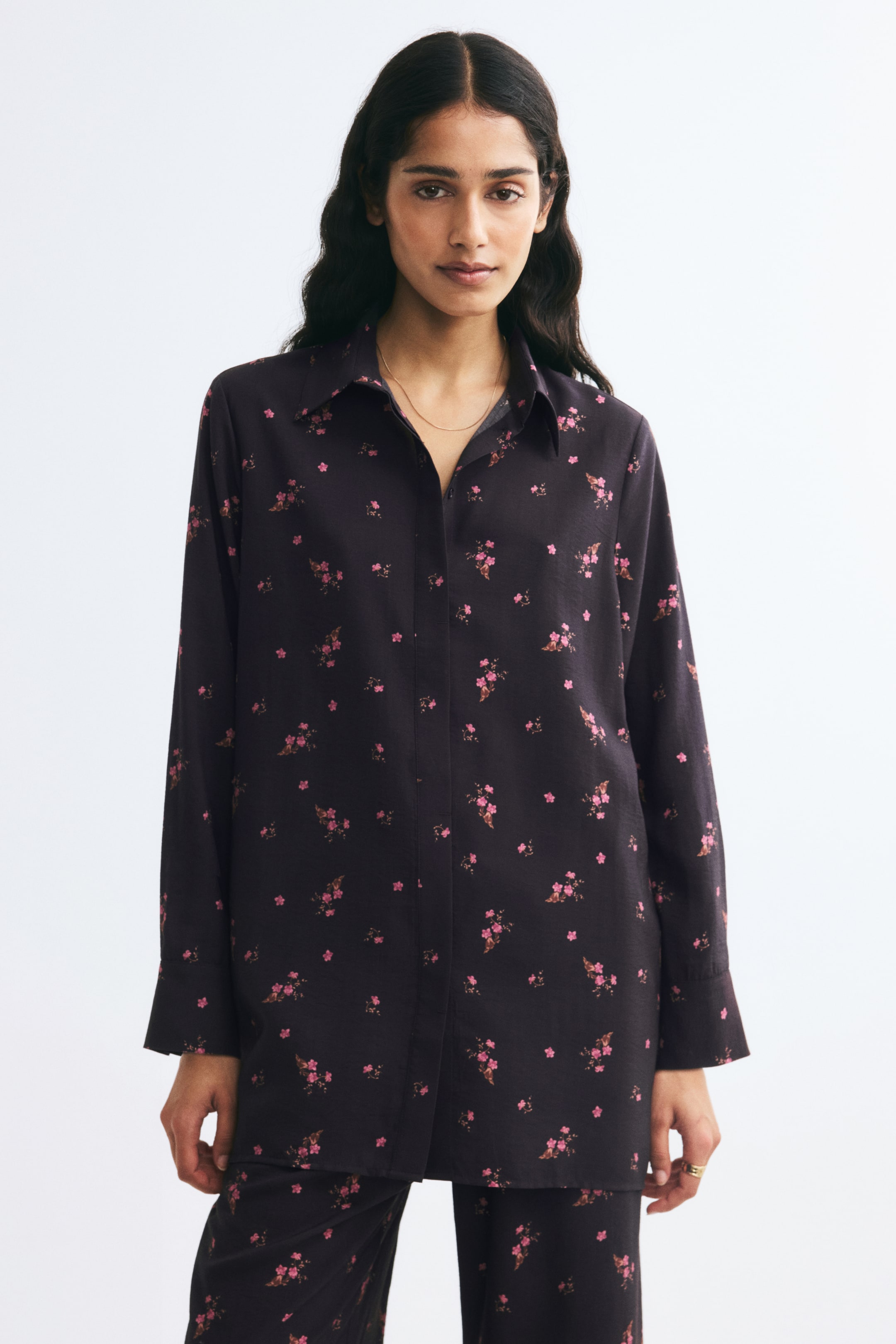 View larger image: A long, loose-fit shirt with a pink floral print is being worn in a front-facing view, featuring a classic collar and a concealed button placket down the front. The shirt drapes loosely with long sleeves ending in button cuffs and features a straight-cut hem.