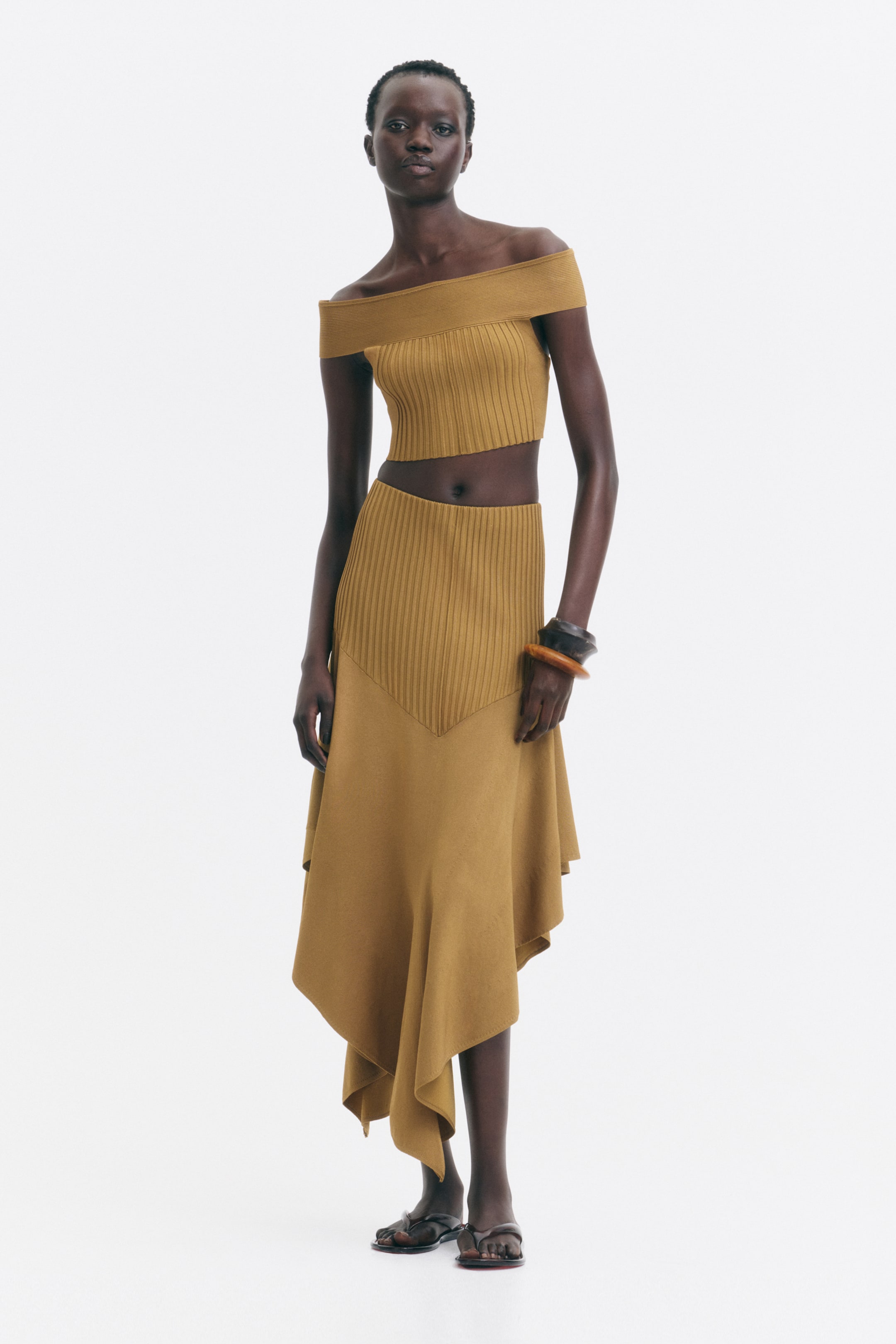 View larger image: The model is wearing a calf-length skirt with a high, fitted rib-knit top section that has a V-shaped bottom edge, flowing into a billowy flounce with an asymmetrical V-shaped hem. It is styled with a matching cropped off-the-shoulder top.