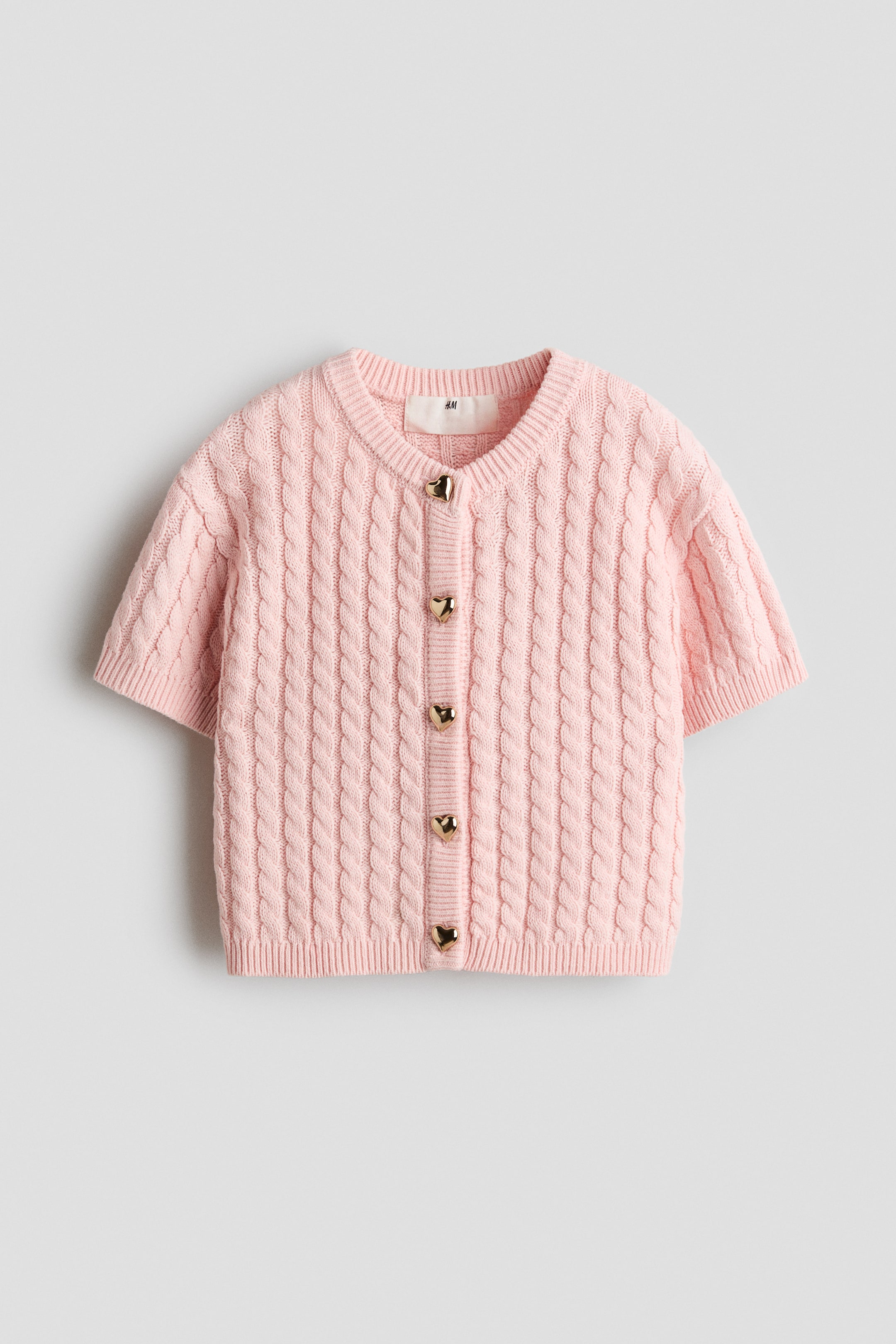 Xem ảnh lớn hơn: Một chiếc áo cardigan tay ngắn màu hồng nhạt đang được chụp từ phía trước, làm nổi bật họa tiết đan cáp phủ toàn bộ bề mặt cùng chi tiết gân ở cổ áo, cổ tay áo và gấu áo. Chiếc cardigan có năm nút cài màu vàng ánh kim hình trái tim dọc theo phần thân trước.