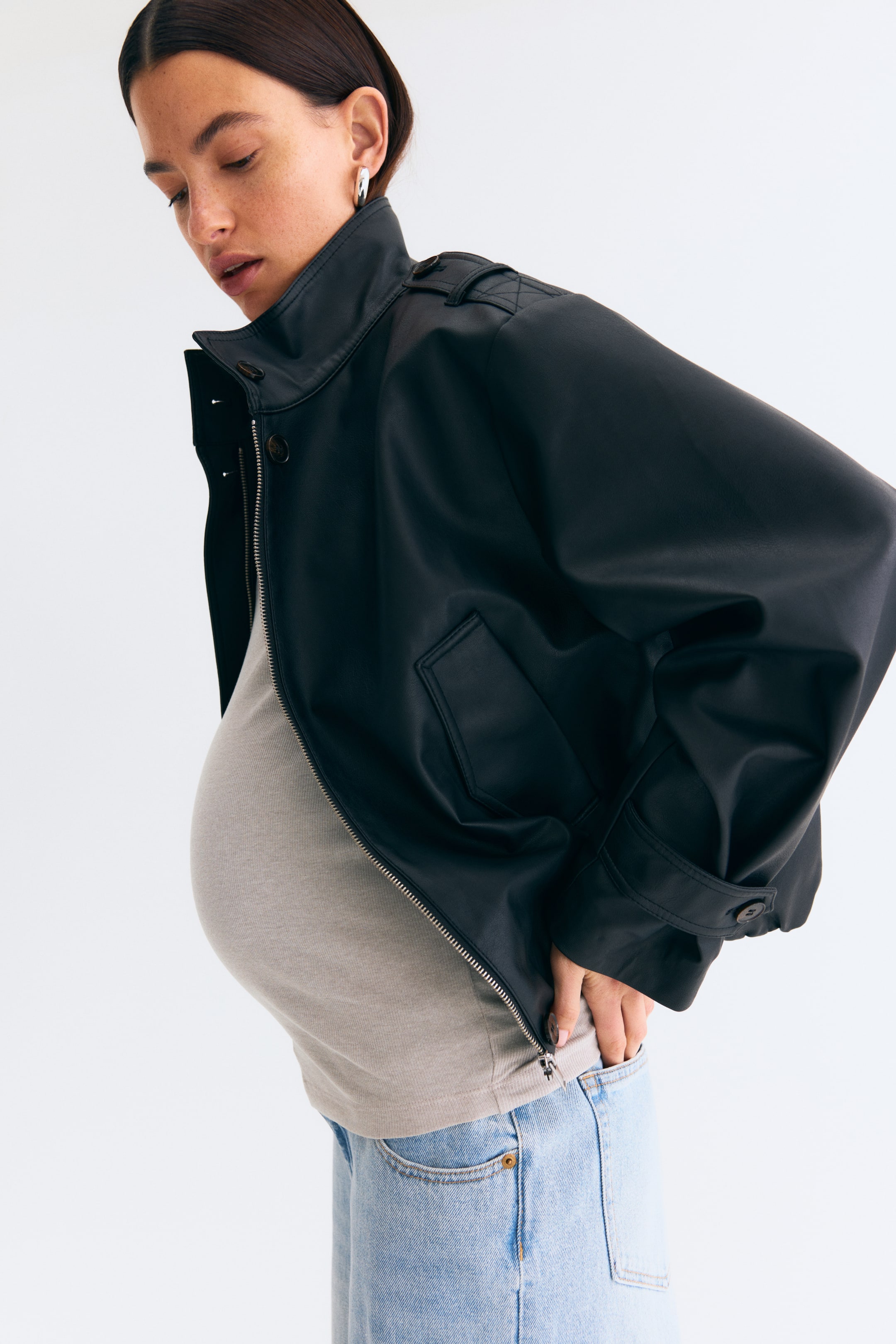 Ver imagen más grande: Se muestra una cazadora bomber de piel sintética negra desde un ligero ángulo lateral-frontal. La cremallera frontal está abierta, dejando ver una camiseta interior gris claro. Un cuello alzado define el escote; charreteras en los hombros y mangas voluminosas que caen con puños abotonados completan la silueta. En la parte inferior delantera se aprecia un bolsillo con solapa.