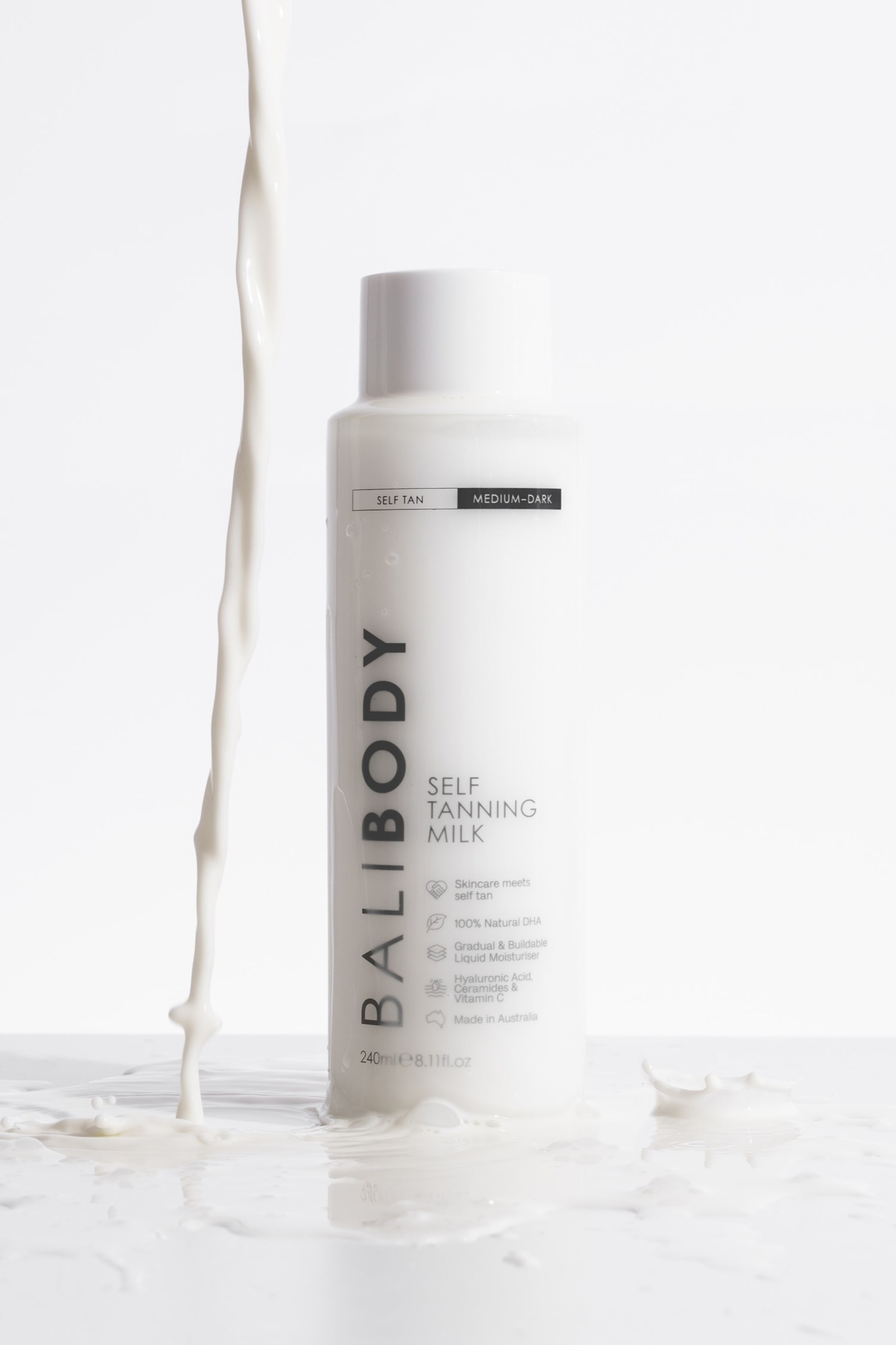 Se større billede: En hvid BALIBODY Self Tanning Milk flaske står oprejst, vendt fremad og lidt til højre, og viser dens frontetiket. Hvid væske hælder ud på venstre side af flasken, og sprøjter omkring dens bund.