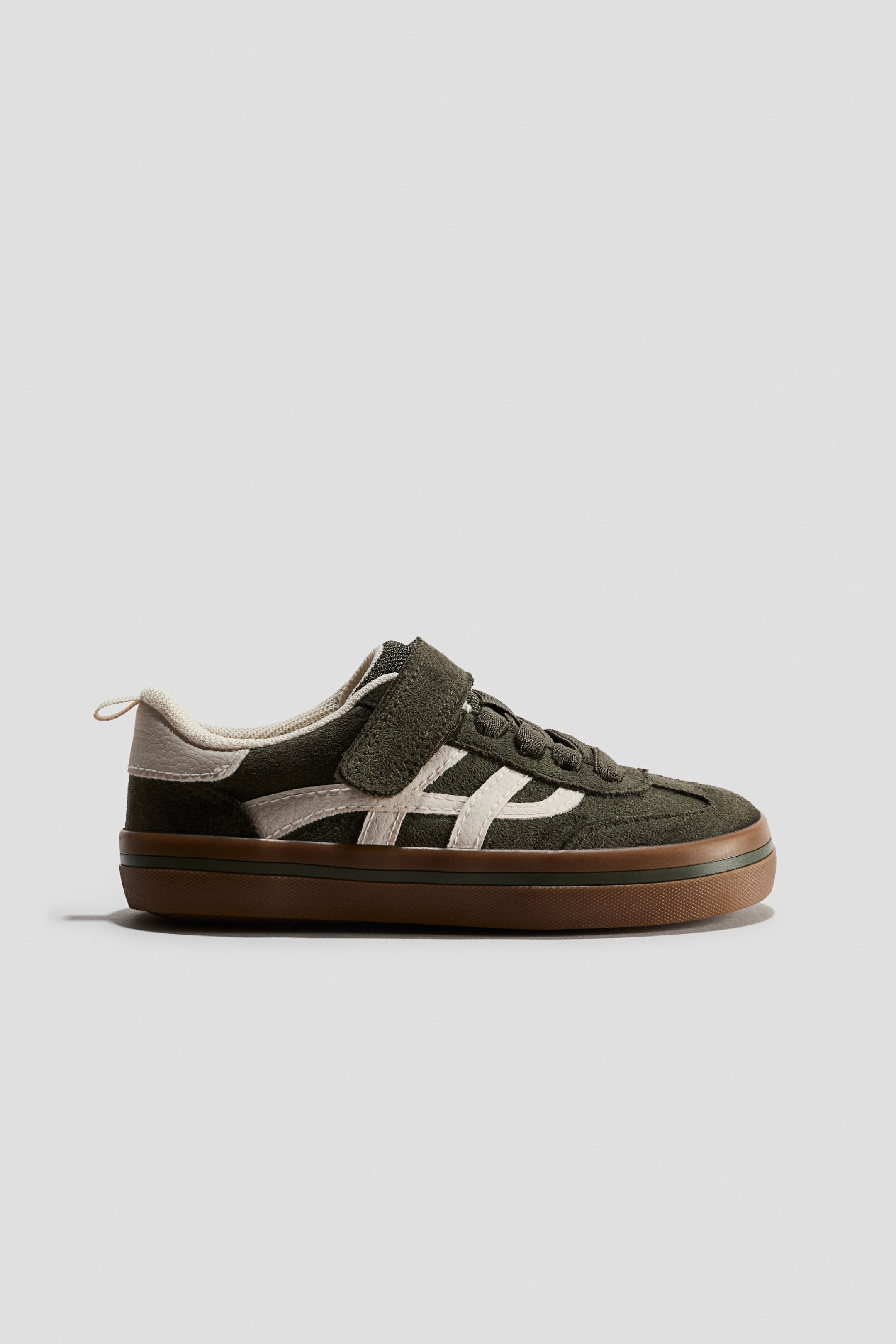 View larger image: A dark green suede low-top sneaker is shown in side profile, featuring a hook-and-loop strap closure and cream-coloured side stripes. The shoe has a thick brown gum sole with a thin green accent line and a cream-coloured heel tab with a pull loop.