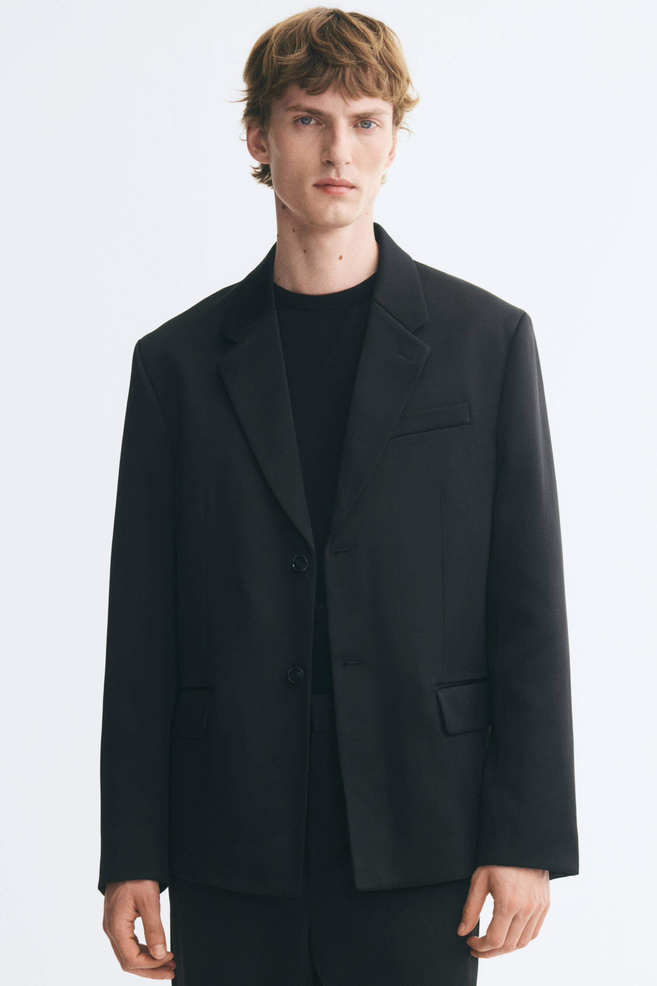 View larger image: The model is wearing a black single-breasted blazer with notched lapels, button fastening, and flap pockets at the waist. It is styled over a matching black crew-neck top and black trousers.