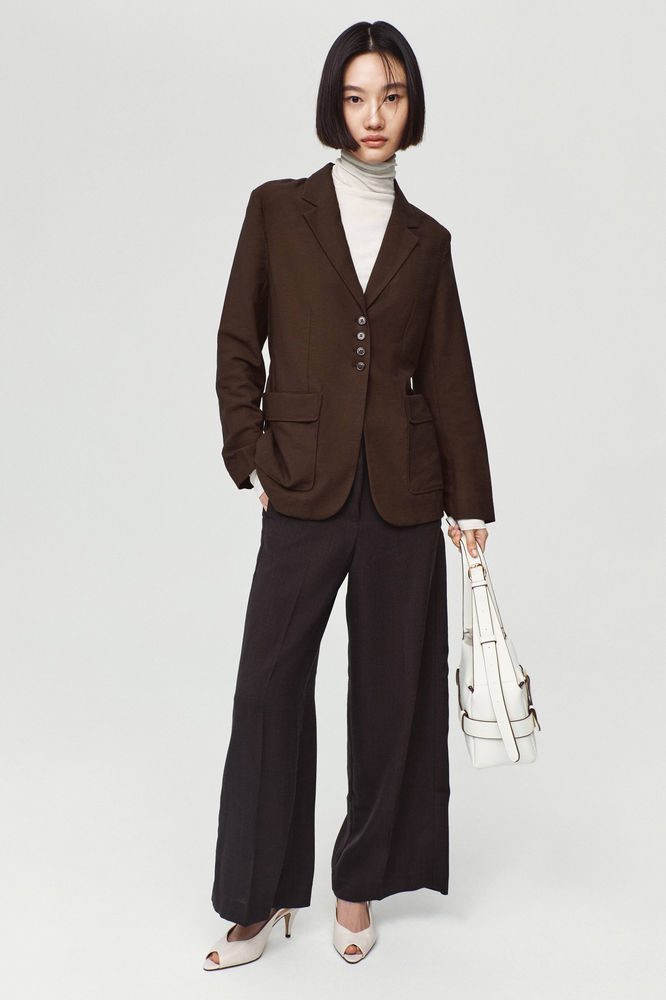 View larger image: A dark brown single-breasted blazer featuring notch lapels, front button closures, and patch pockets with flaps is worn by the model. The blazer is layered over a cream turtleneck top and paired with dark, wide-leg trousers.