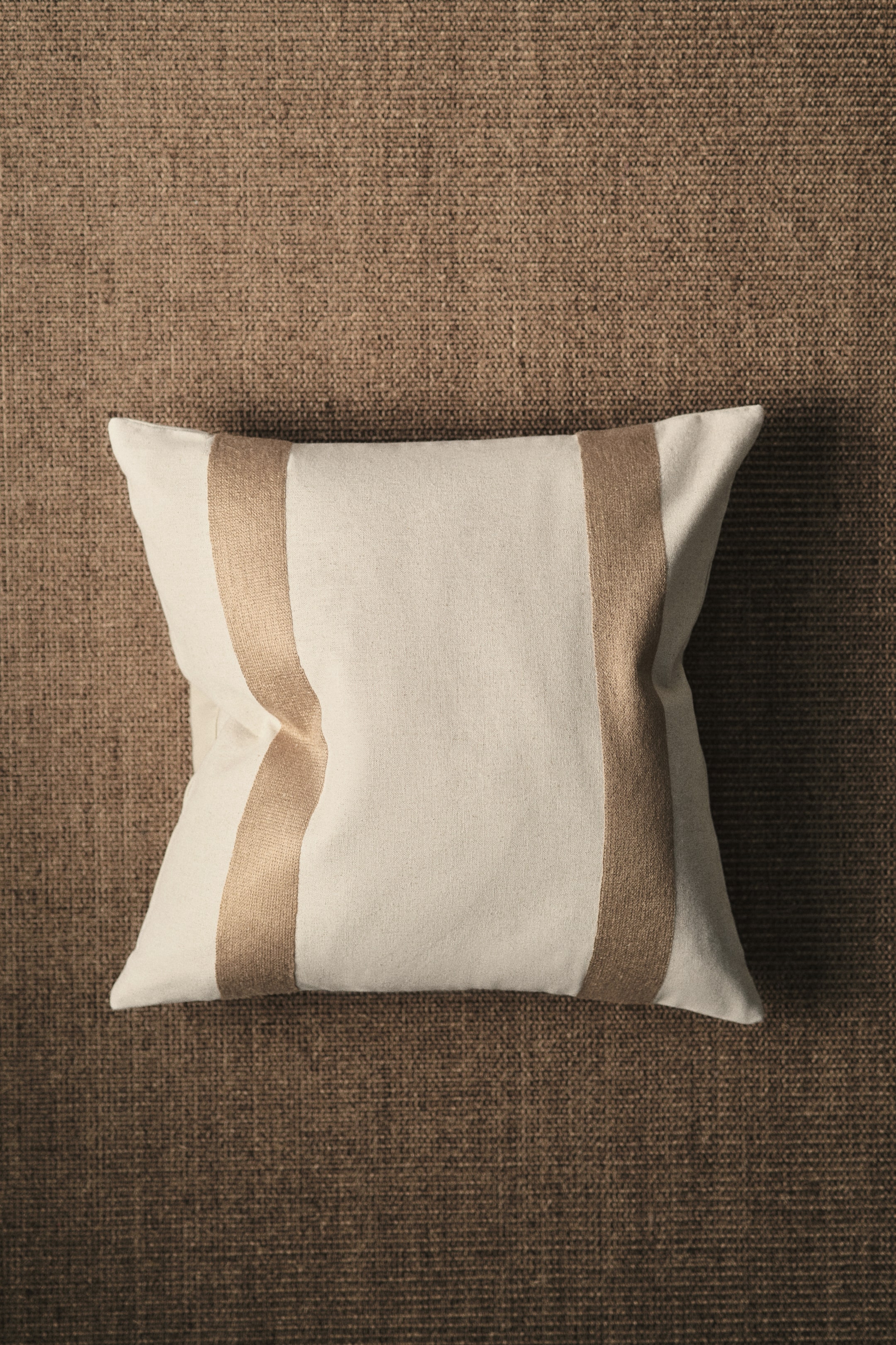 View larger image: The cushion cover showcases its front, crafted from a rustic linen and cotton blend fabric in an off-white hue. It features broad, vertically embroidered stripes in a warm brown shade, with a zip closure along one side.