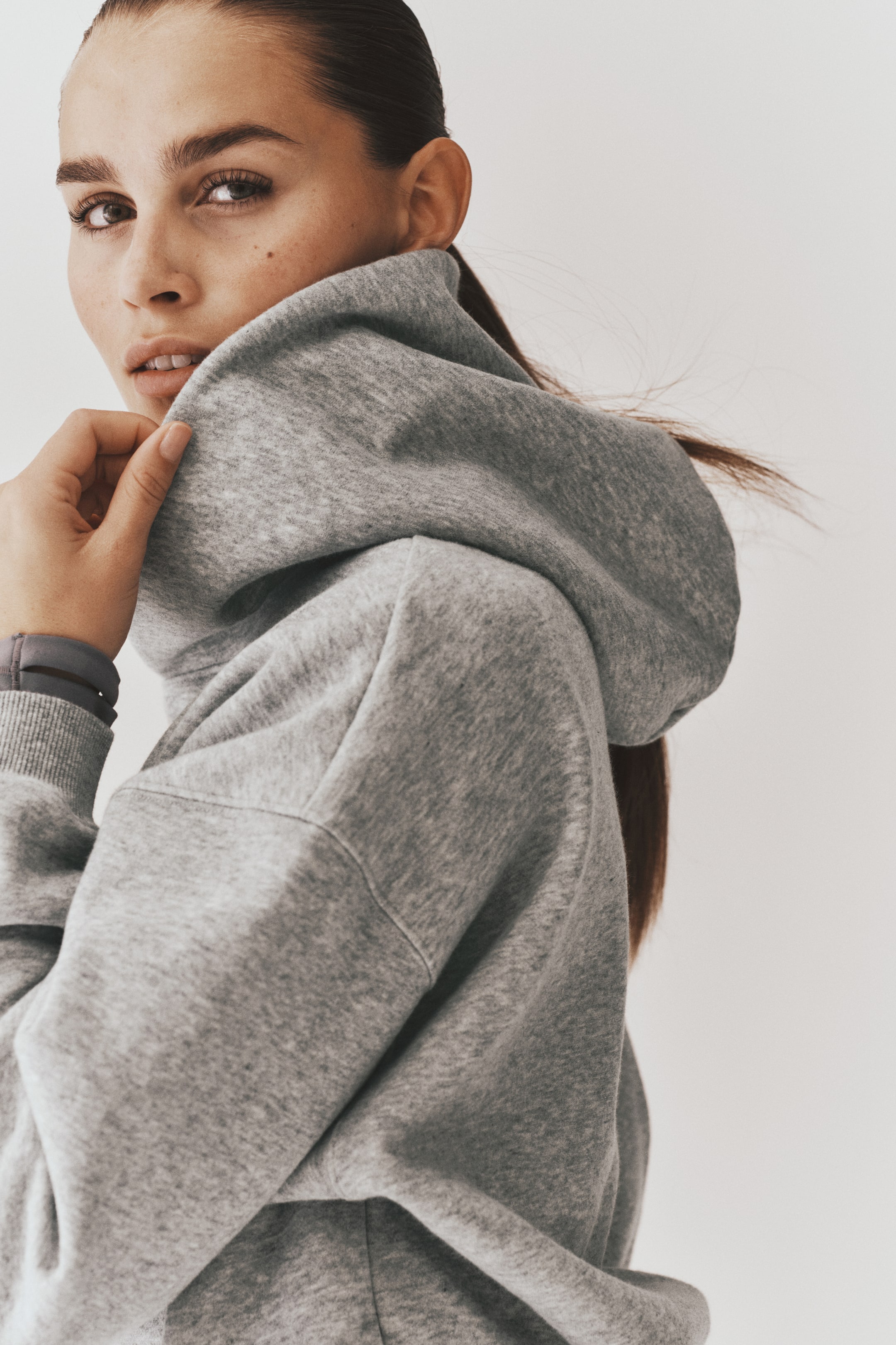 Ampliar la imagen: La modelo lleva puesta una sudadera con capucha de forro polar gris de corte oversize, con capucha cruzada de doble capa y hombros caídos. La modelo sostiene el costado de la capucha gruesa cerca de su rostro, mostrando el puño acanalado de la manga.