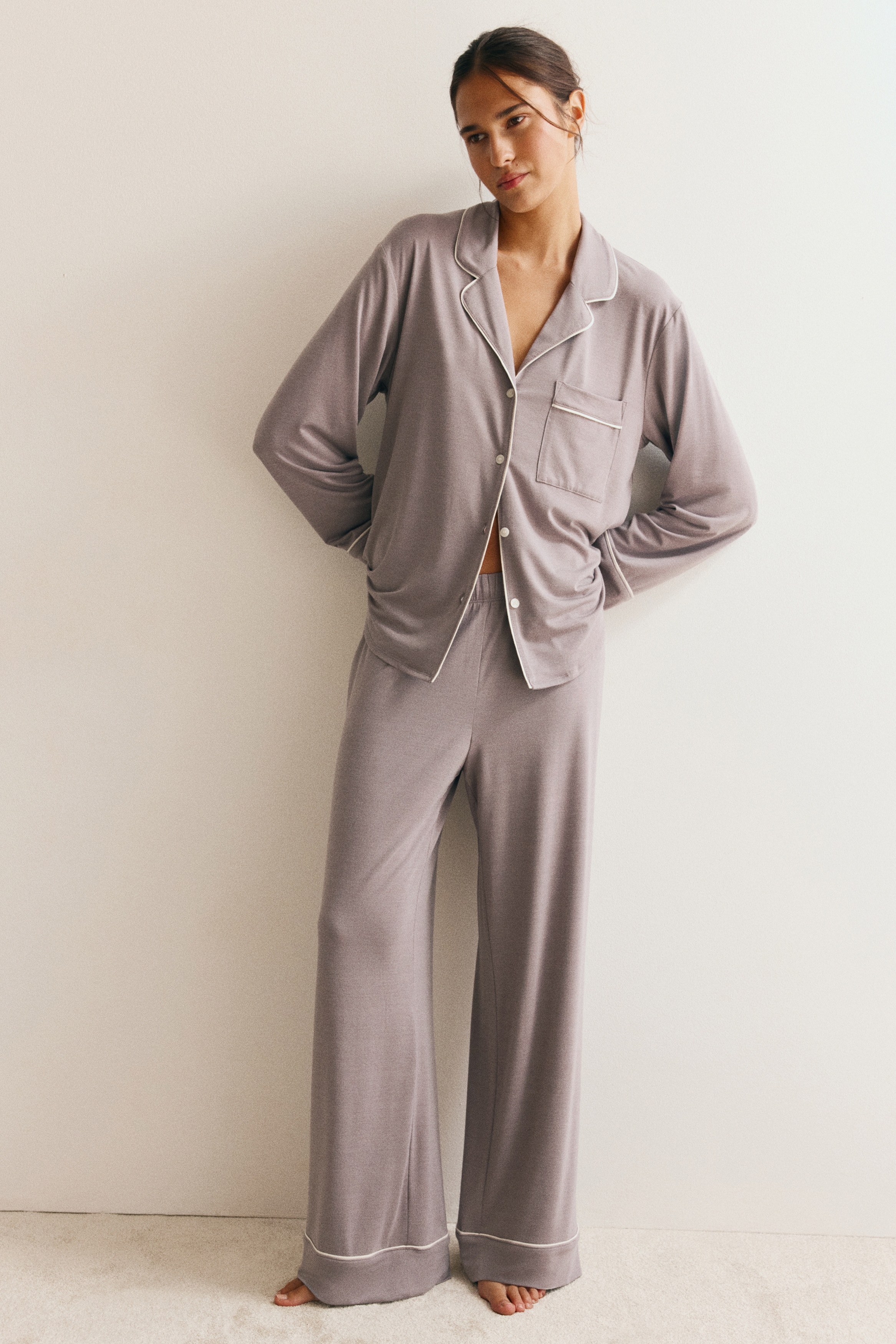 Ein grauviolettes Jersey-Pyjamaset hÃ¤ngt in frontaler Ansicht und besteht aus einem Langarmshirt und einer dazu passenden Hose mit weitem Bein. Beide Teile sind mit feiner weiÃer Kontrastpaspelierung entlang des Kragens, der Brusttasche, der ÃrmelbÃ¼ndchen und der Hosenbeine versehen.