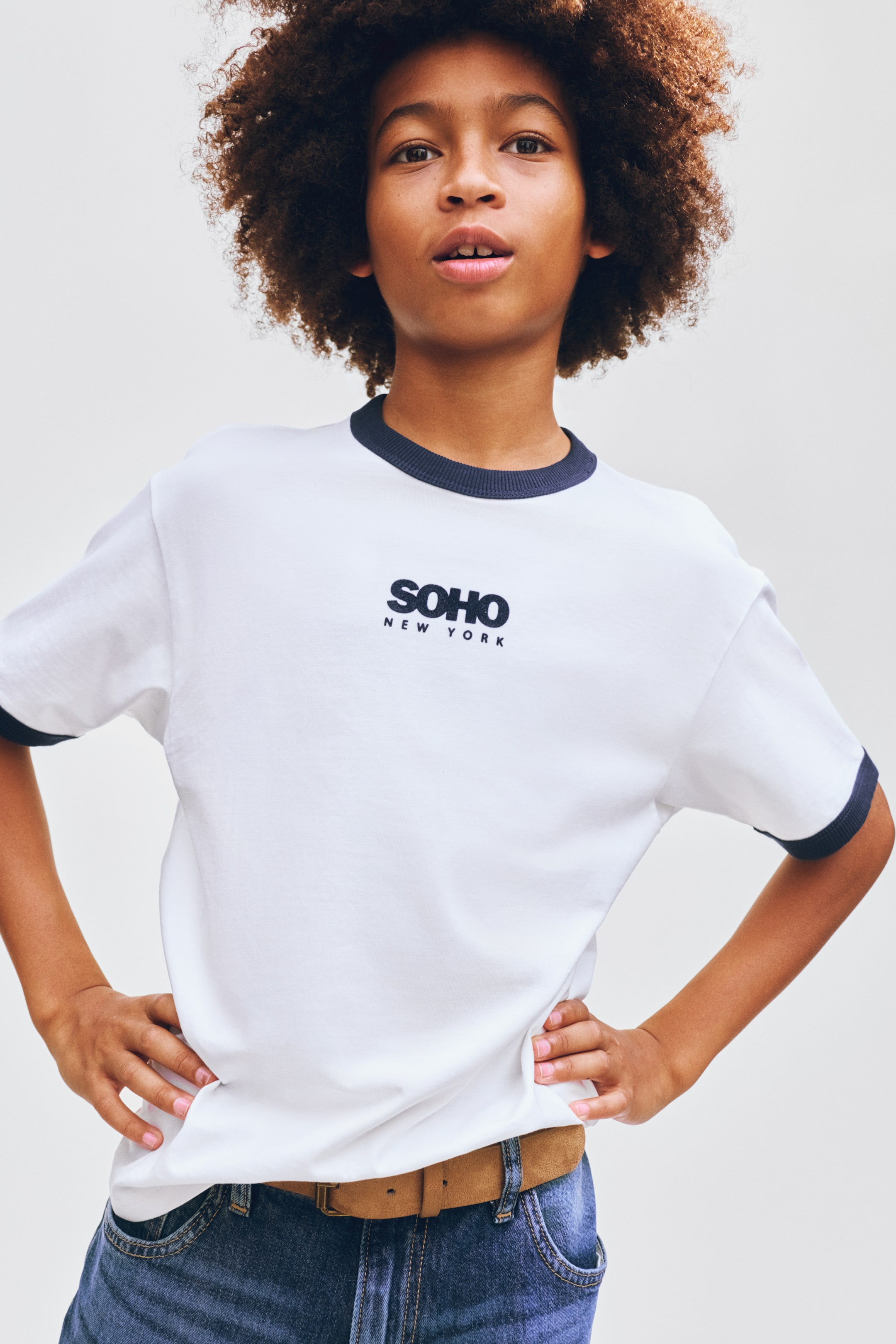 Größeres Bild anzeigen: Ein weißes Ringer-T-Shirt mit einem marineblauen gerippten Rundhalskragen und passenden gerippten kurzen Ärmelbündchen, mit "SOHO" in kräftigen marineblauen Buchstaben und "NEW YORK" darunter auf der Brustmitte.