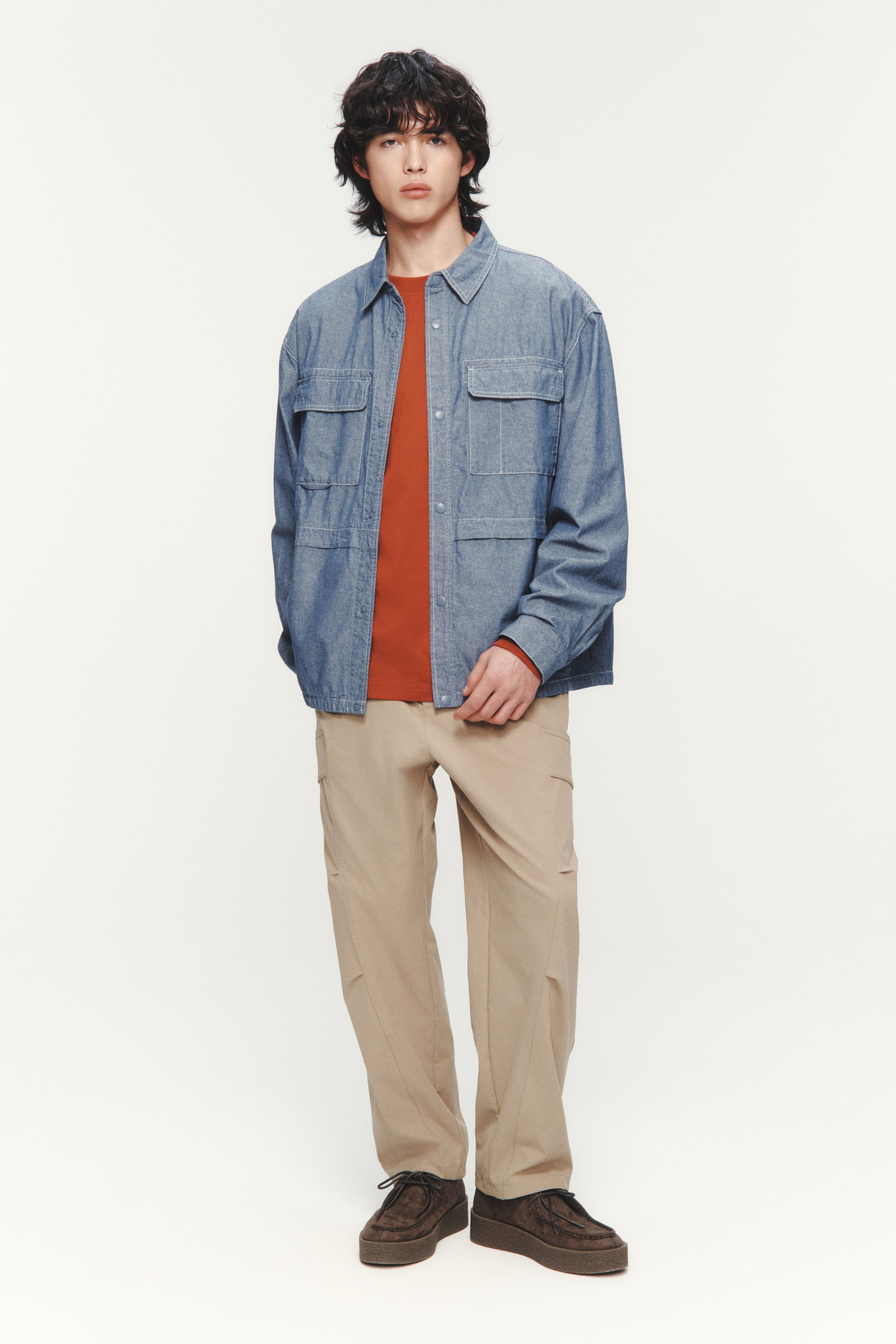 이미지 확대 보기: 이 제품은 뾰족한 칼라와 네 개의 플랩 포켓이 있는 연한 파란색 샴브레이 오버셔츠를 특징으로 하며, 녹슨 색상의 긴 소매 크루넥 셔츠 위에 오픈 스타일로 착용되었습니다. 연한 베이지색 카고 팬츠와 두꺼운 밑창을 가진 짙은 갈색 스웨이드 레이스업 슈즈로 완성되었습니다.