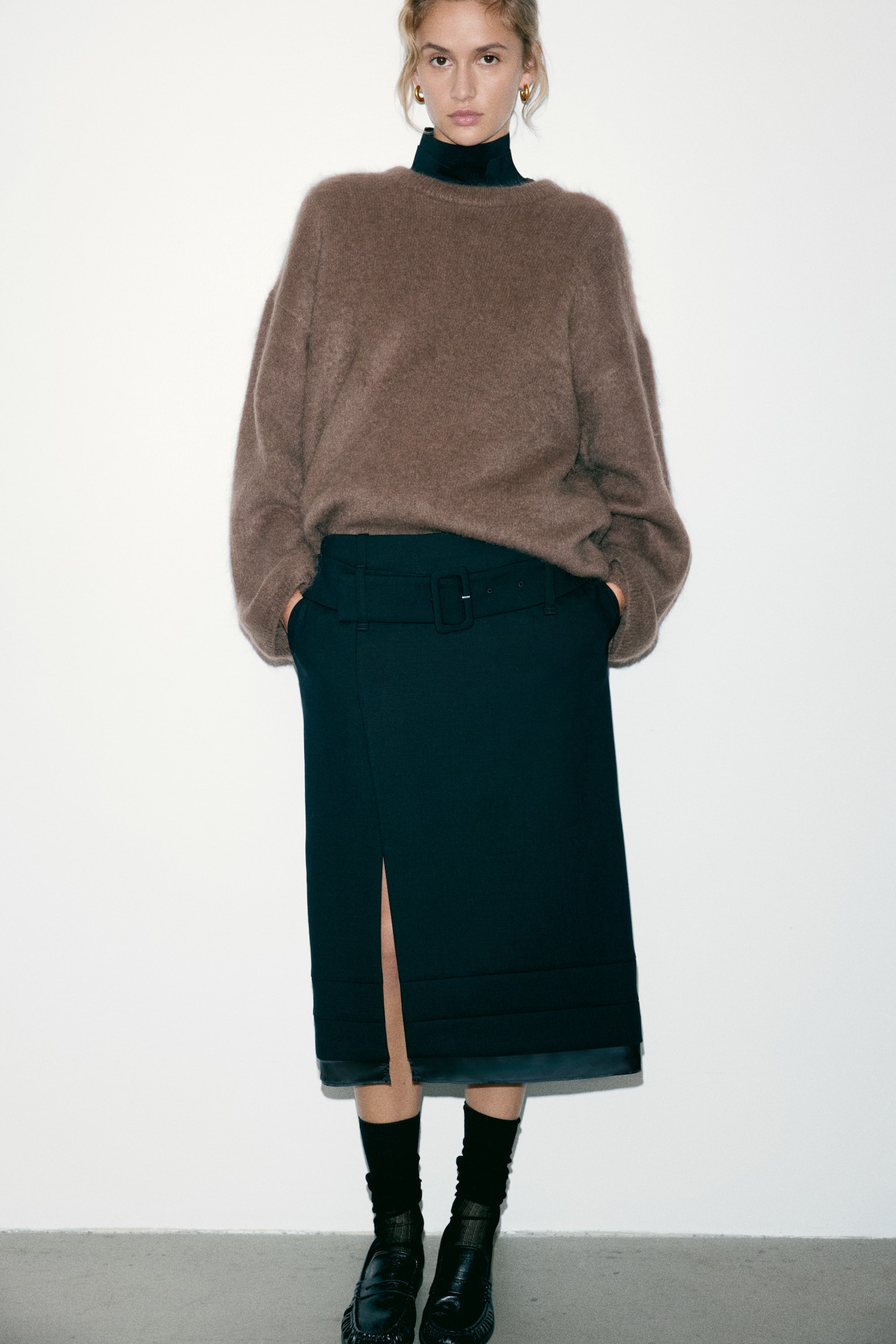 An oversized brown knit jumper featuring dropped shoulders and a soft texture is being worn by the model over a dark high-neck top. The jumper, which has ribbing at the neckline, cuffs, and hem, is being styled with a dark, belted midi skirt. - 1