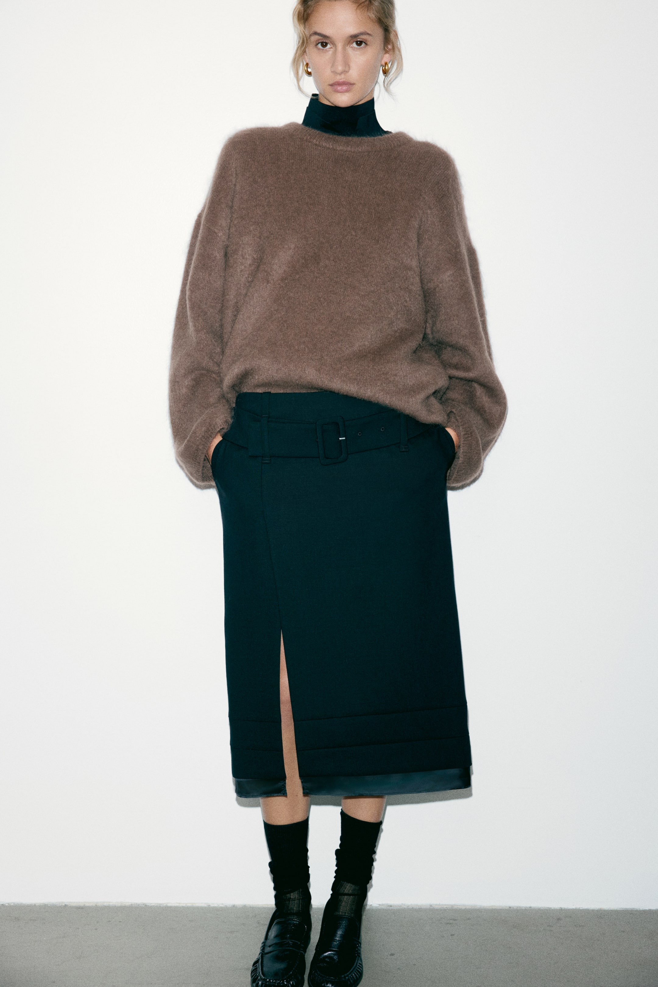 Agrandir l'image: Un pull en maille marron oversize, à épaules tombantes et à texture douce, est porté par le mannequin par-dessus un haut foncé à col montant. Le pull, doté de bords-côtes au niveau de l’encolure, des poignets et de l’ourlet, est associé à une jupe midi foncée ceinturée.