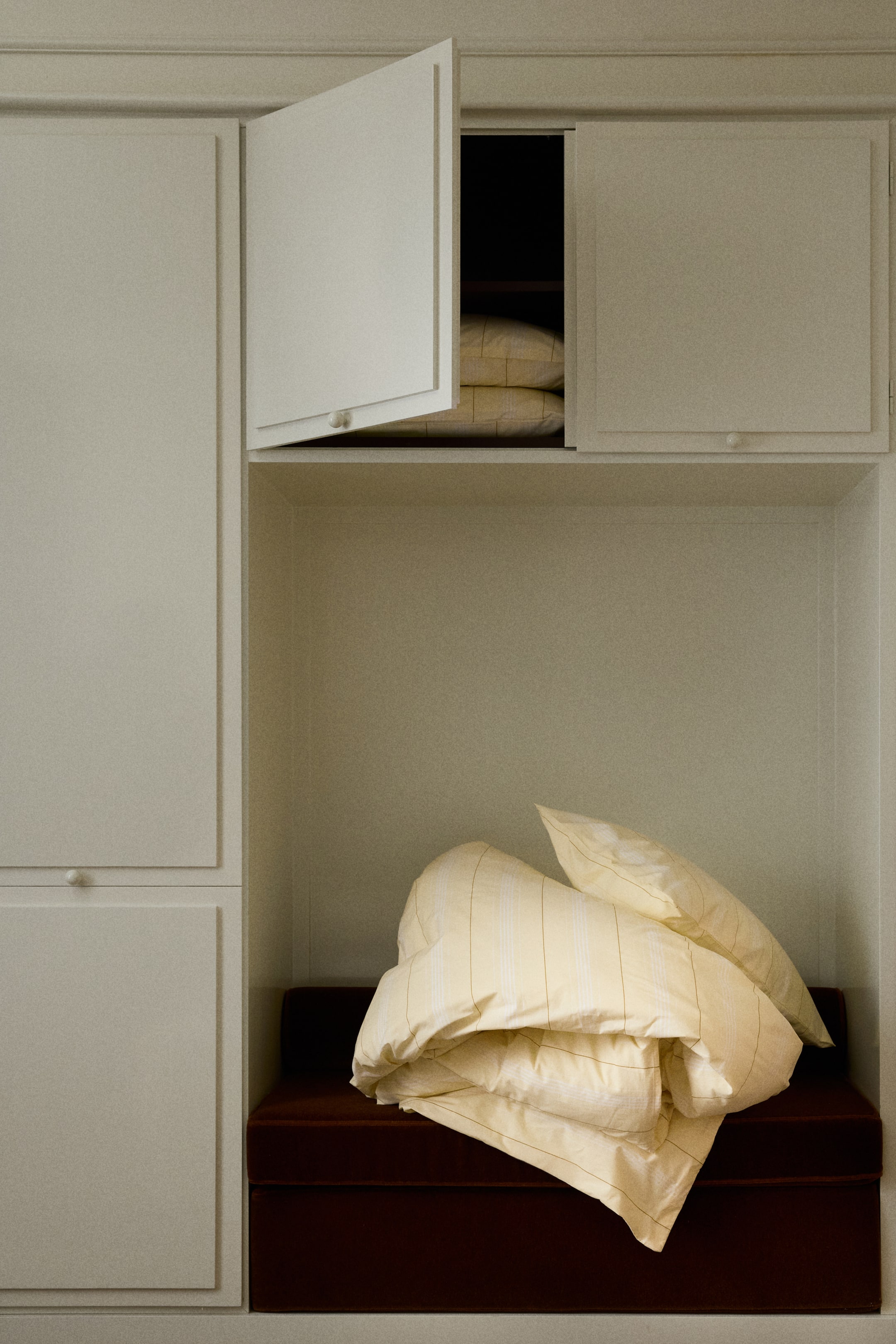 View larger image: A light yellow duvet and two matching pillows, featuring thin vertical stripes, are folded and casually arranged on a dark brown cushion.