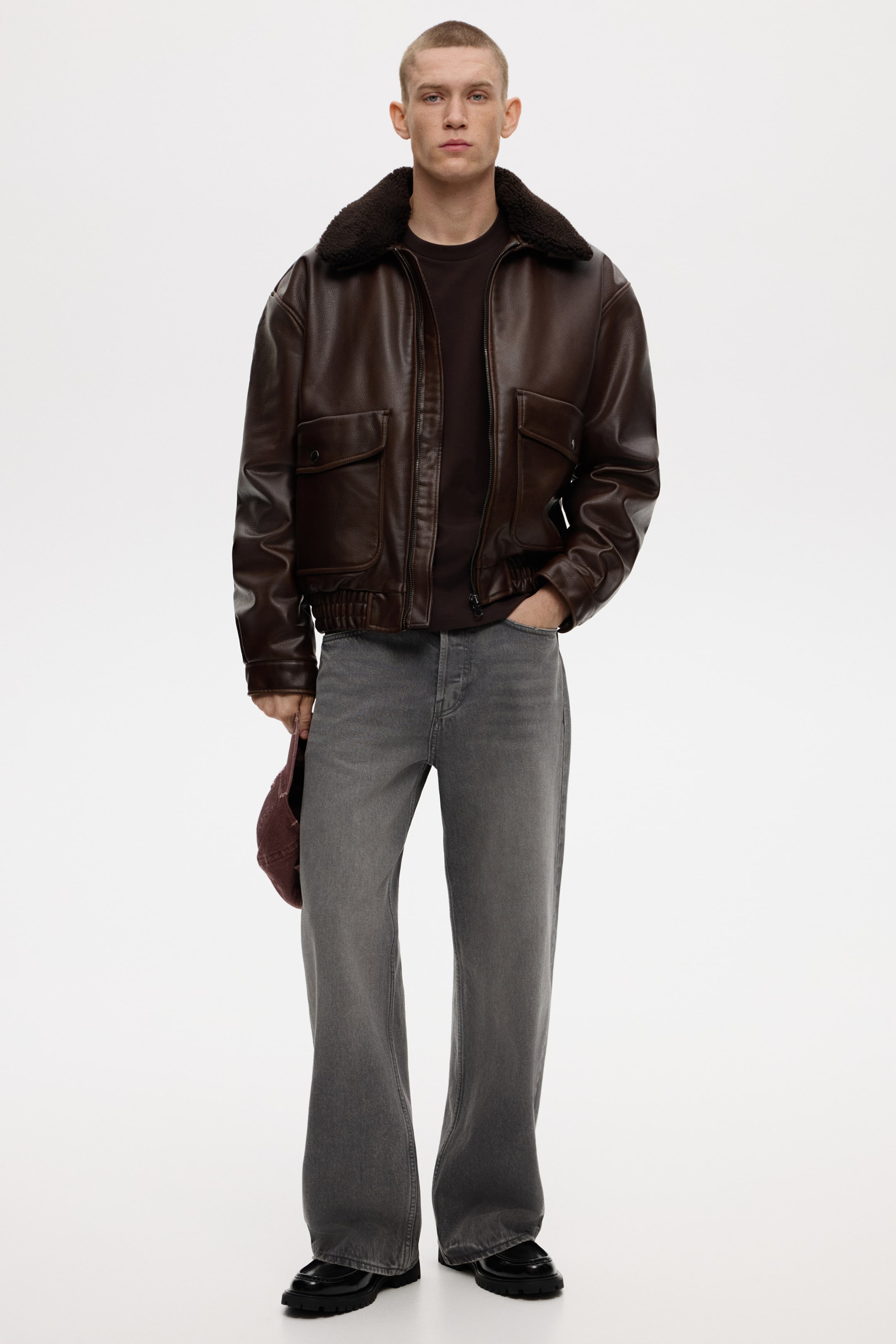 View larger image: A dark brown bomber jacket features a smooth, slightly shiny finish and a large, textured dark brown collar. The jacket is designed with two flap pockets, elasticated cuffs and an elasticated hem, and is worn over a brown T-shirt.