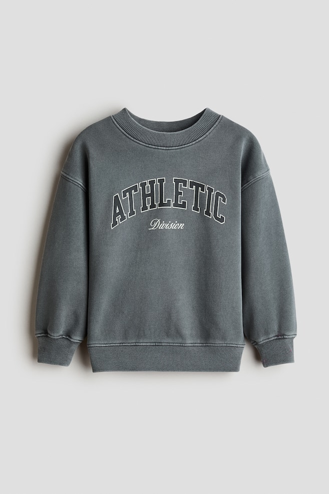 A front-facing view shows a loose-fit, washed dark grey sweatshirt featuring a round, rib-trimmed neckline, dropped shoulders, and ribbed cuffs and hem. The front displays the words "ATHLETIC Division" in an arched, white-outlined varsity font. - 1