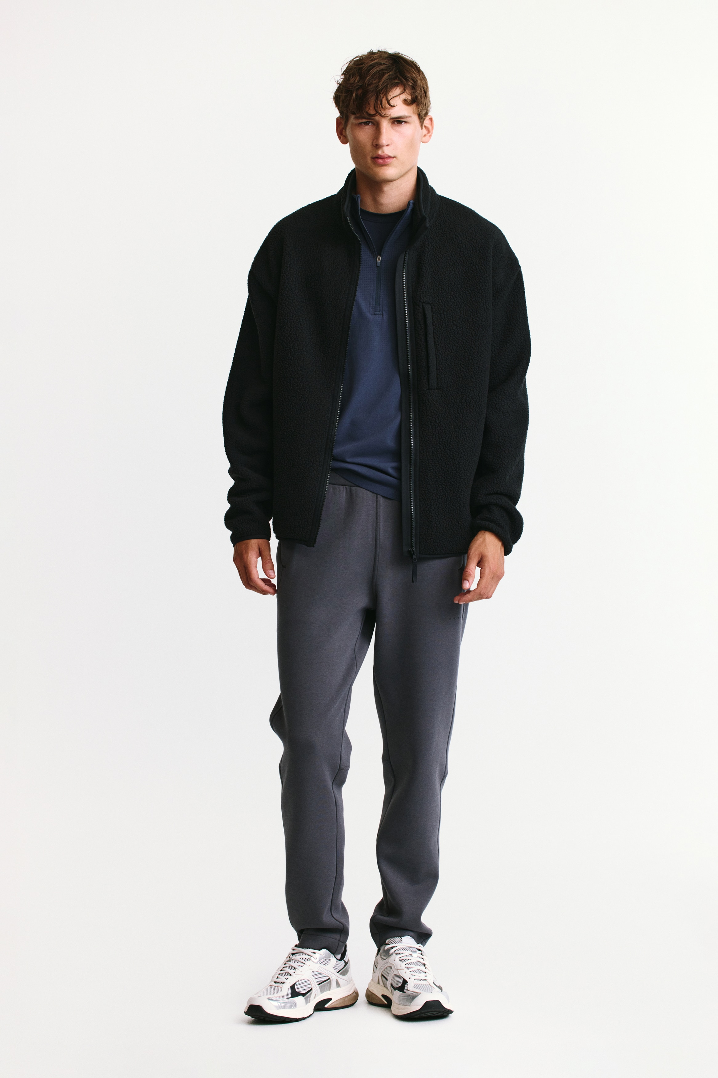 A pair of dark grey joggers is shown front-facing, featuring an elasticated waistband, vertical zippered side pockets, and tapered legs. The trousers are made from a smooth, structured fabric with seam detailing near the knees and clean-finished hems.